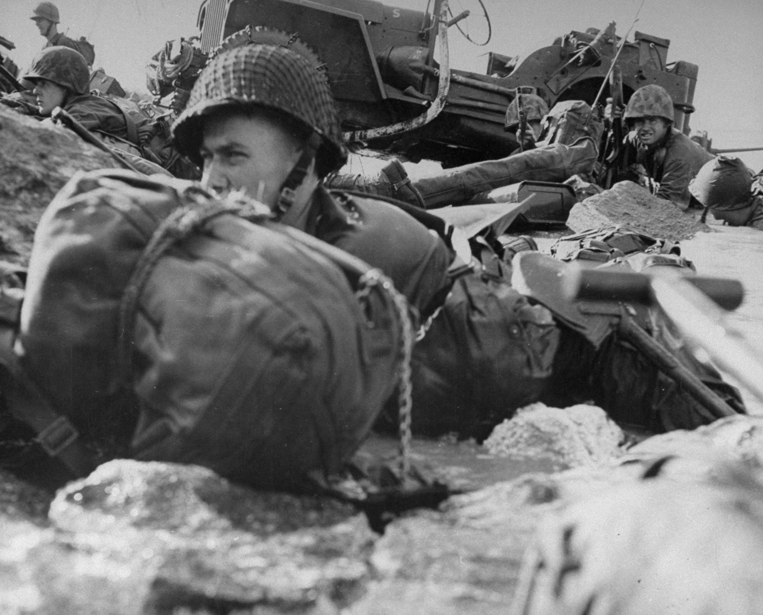 Marines hug sharp coral of beach, with pillbox only 12 yards in front of them. Strock, still lying in the water, has poked camera just over low pile of coral. Men finally edged back into the surf and moved cautiously down beach to flank pillbox. Marine in the background at upper left shows dangerous curiosity.