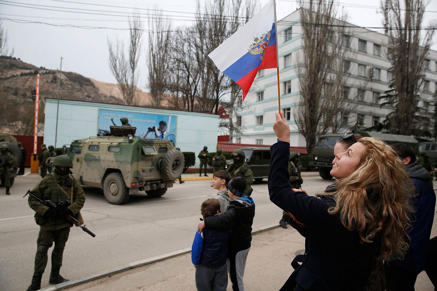 A woman waves a Russian flag as armed servicemen wait near Russian army vehicles outside a Ukrainian border guard post in the Crimean town of Balaclava