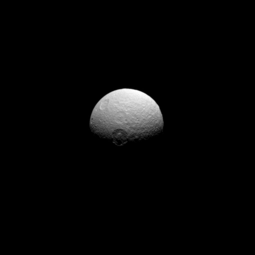 Tethys, a mid-sized moon of Saturn, hangs seemingly motionless and alone in space. The craters Melanthius (near the center), Dolius (above Melanthius), and Penelope (upper left, almost over the edge of the moon) are visible.