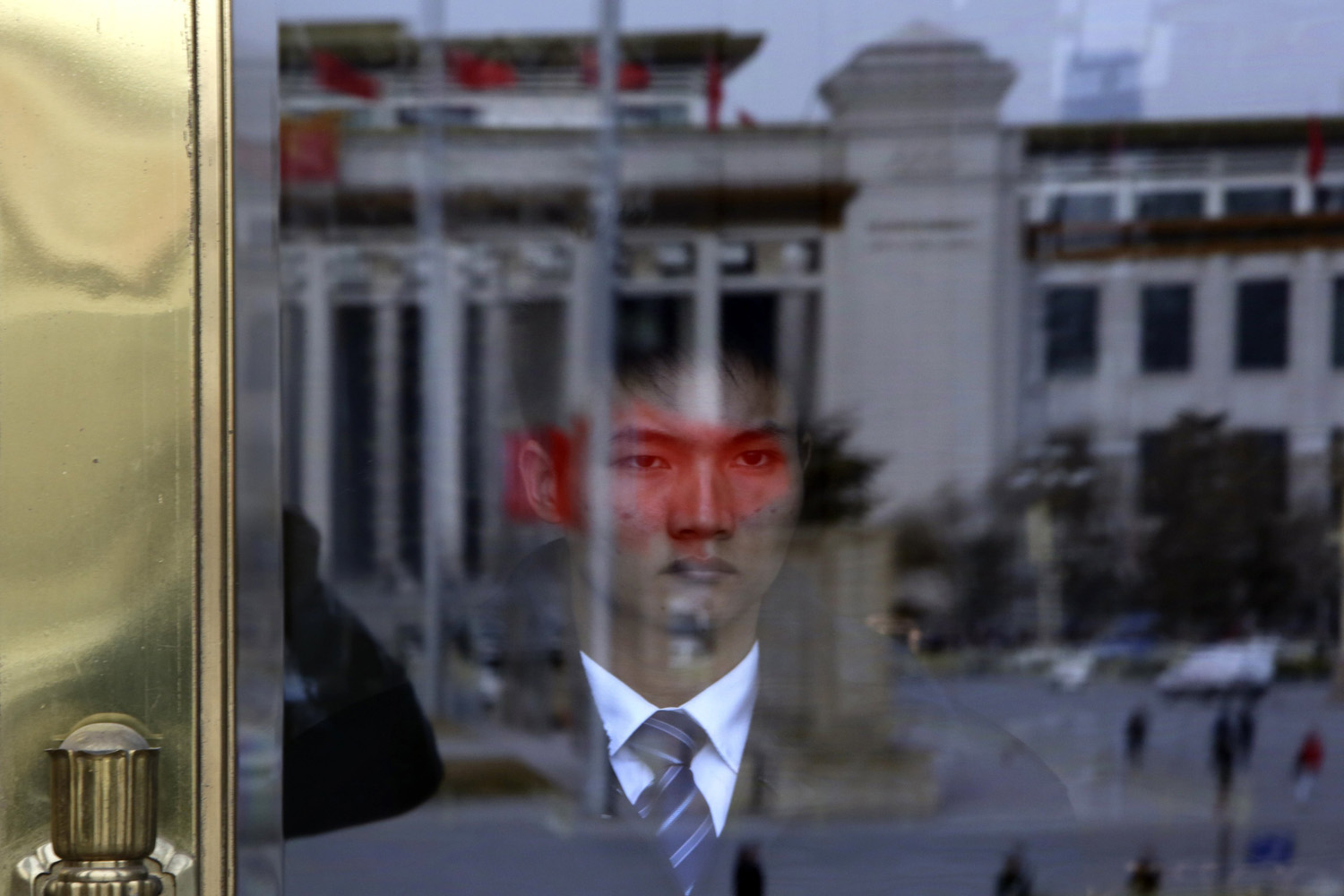 A soldier in plain clothes from the Chinese PLA stands guard behind an entrance of the Great Hall of the People as red flags are reflected on a glass door in Beijing