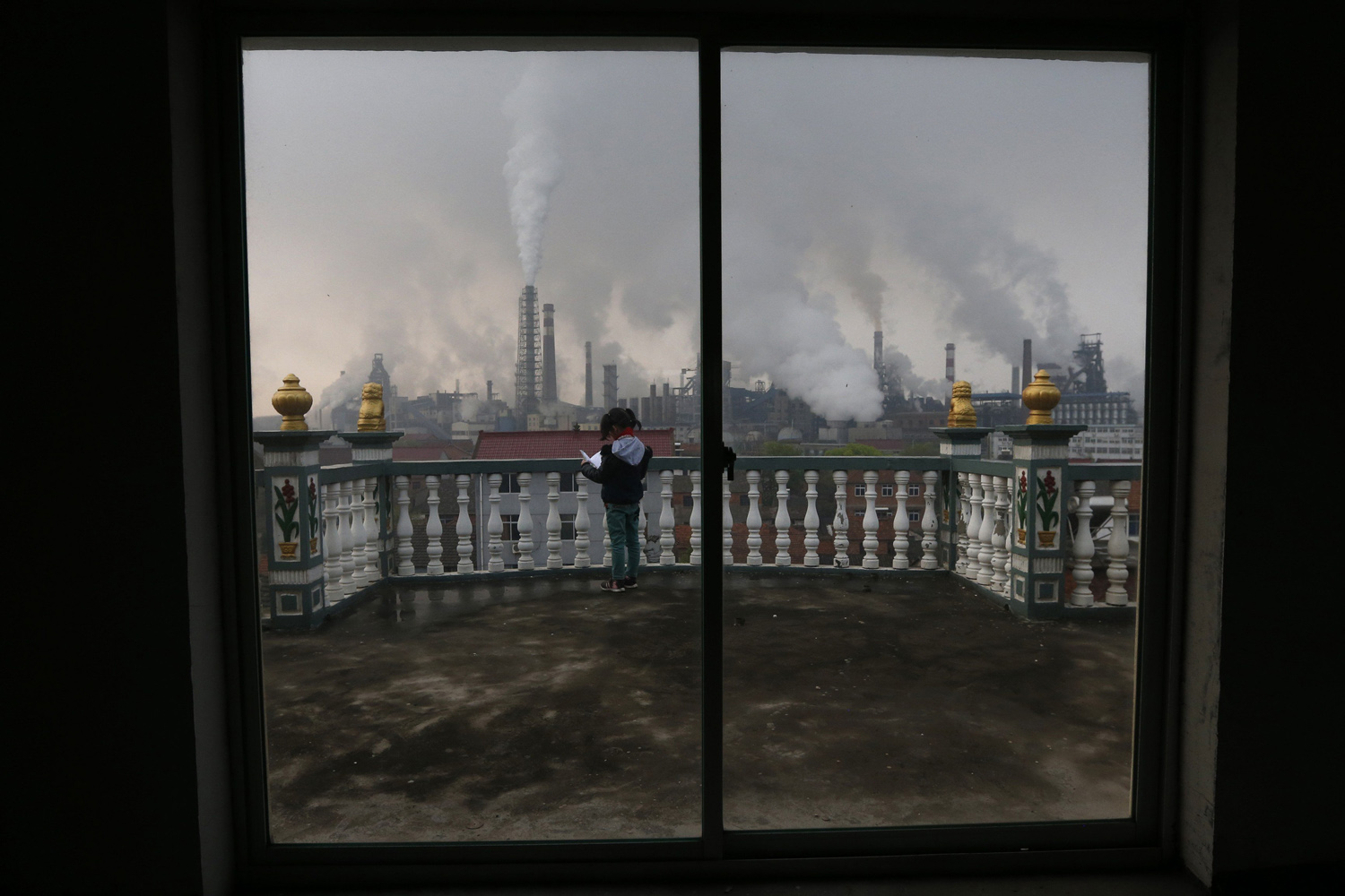 A girl reads a book on her balcony as smoke rises from chimneys of a steel plant, on a hazy day in Quzhou