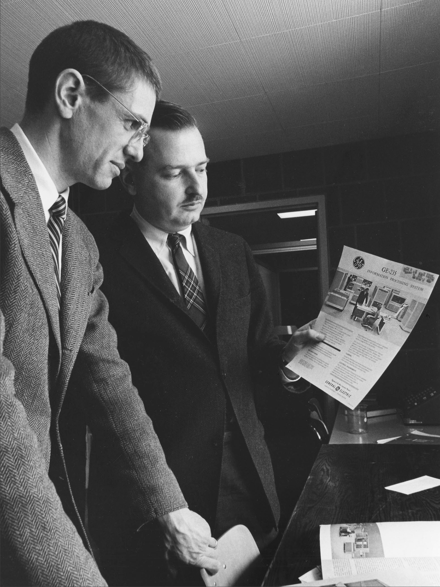 Tom Kurtz and John Kemeny examine a brochure for the GE-225 mainframe computer that powered the initial version of Dartmouth's time-sharing system (Adrian N. Bouchard / Dartmouth College)