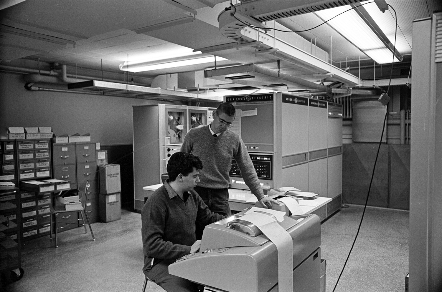 Tom Kurtz (standing) works with Michael Busch, co-programmer of the DTSS, with the GE-225 mainframe in the background (Adrian N. Bouchard / Dartmouth College)