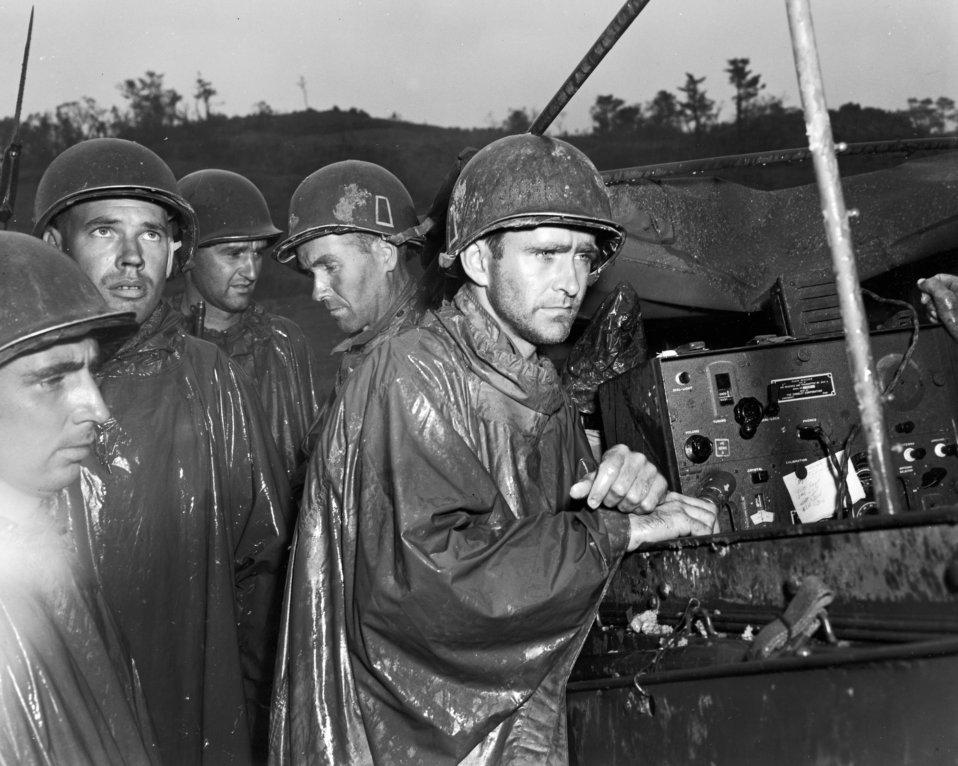 Grim-faced American soldiers fighting on Okinawa listen to a radio broadcast of the surrender of Germany and the end of WWII in Europe, May 1945.