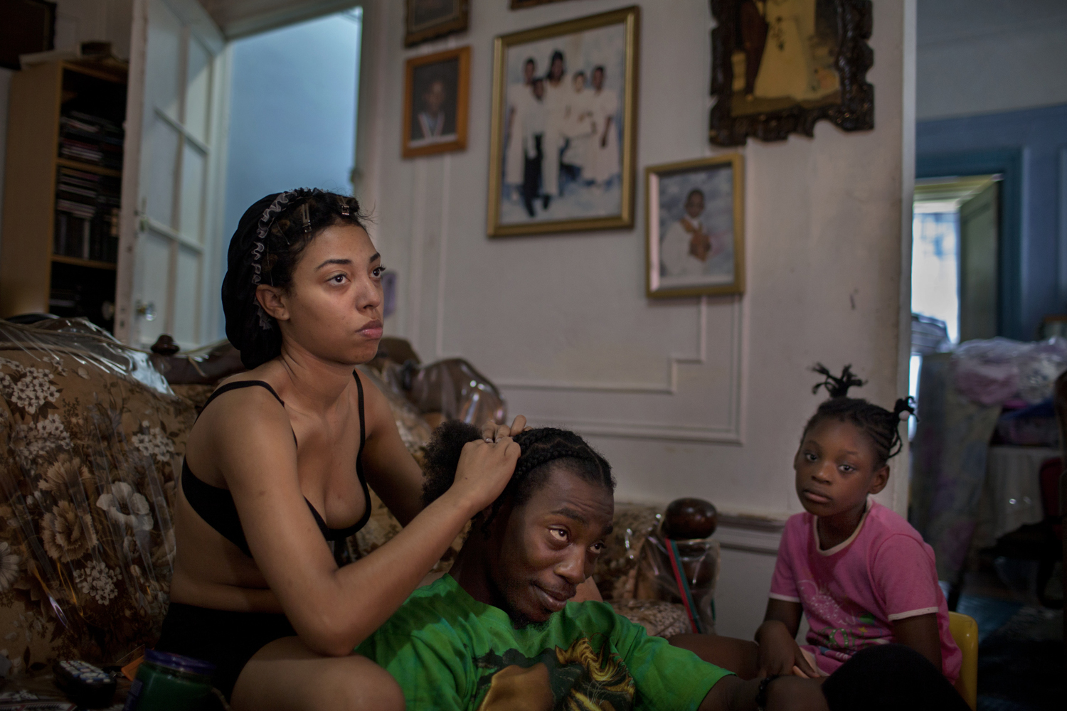 August 8, 2013. East Flatbush, Brooklyn. Sarah braids her brother's hair in their mother's home as her niece looks on.