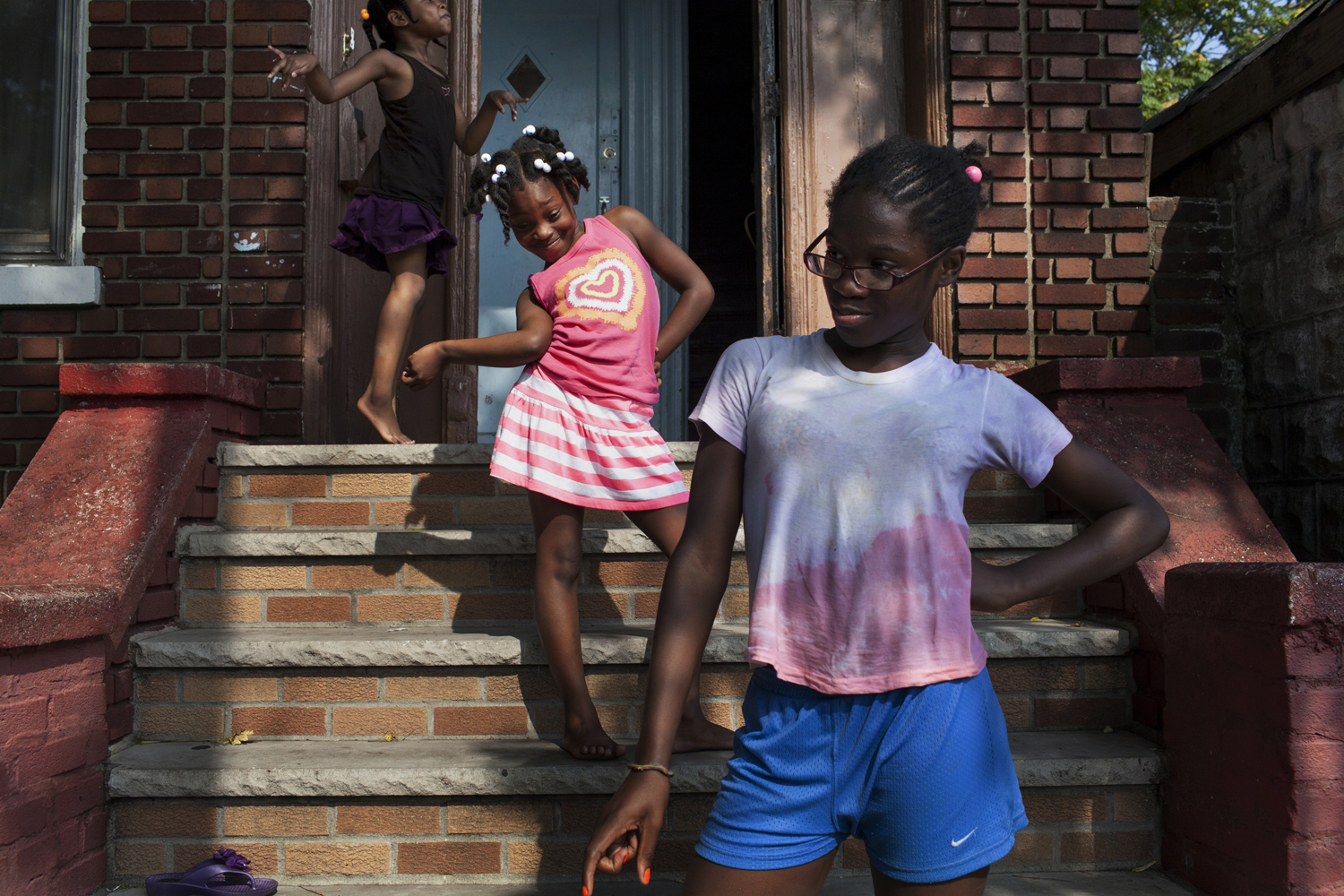August 21, 2013. Flatbush, Brooklyn. Sarah's niece and her friends strike poses on the stoop of Sarah's home on a summer afternoon. Dancehall dancers often site their dance abilities as part of their Caribbean heritage. Young girls start to learn some of the less suggestive dance moves in a dance hall queen's repertoire very young, but Sarah's niece giggle and exclaims "We're too young!" when asked if she can dance like her auntie. Two months later, Sarah will be shot in the foot just a few feet from this stoop.(Natalie Keyssar)