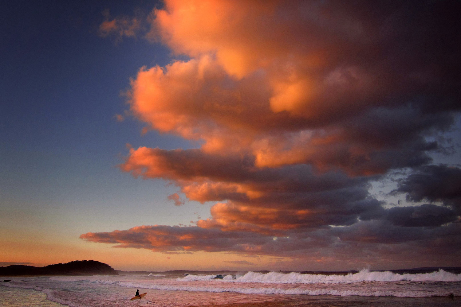 Surfer holding her board wades through the surf as clouds above are lit by the setting sun at Mollymook Beach on the south coast of New South Wales