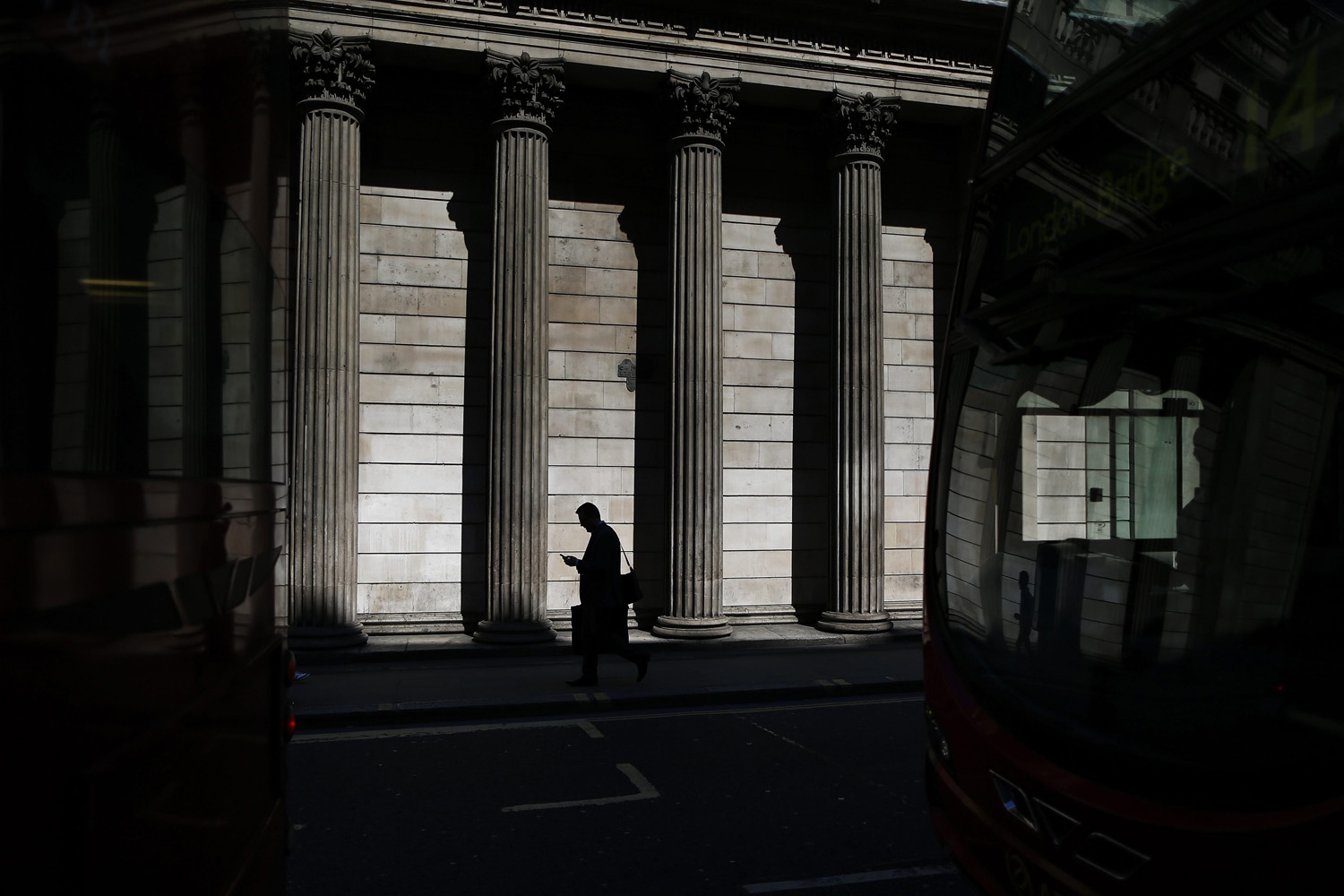 A man is reflected in a bus windscreen as he walks past the columns of the Bank of England on a sunny morning in the City of London