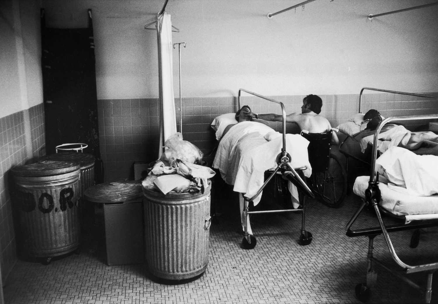 At the Bronx VA Hospital veteran Frank Stoppiello, wounded in the Ashau Valley in Vietnam, gives a cigarette to quadriplegic Andrew Kmetz, an Army veteran, as they wait for treatment. Because of overcrowding, they must share a corner with trash cans.