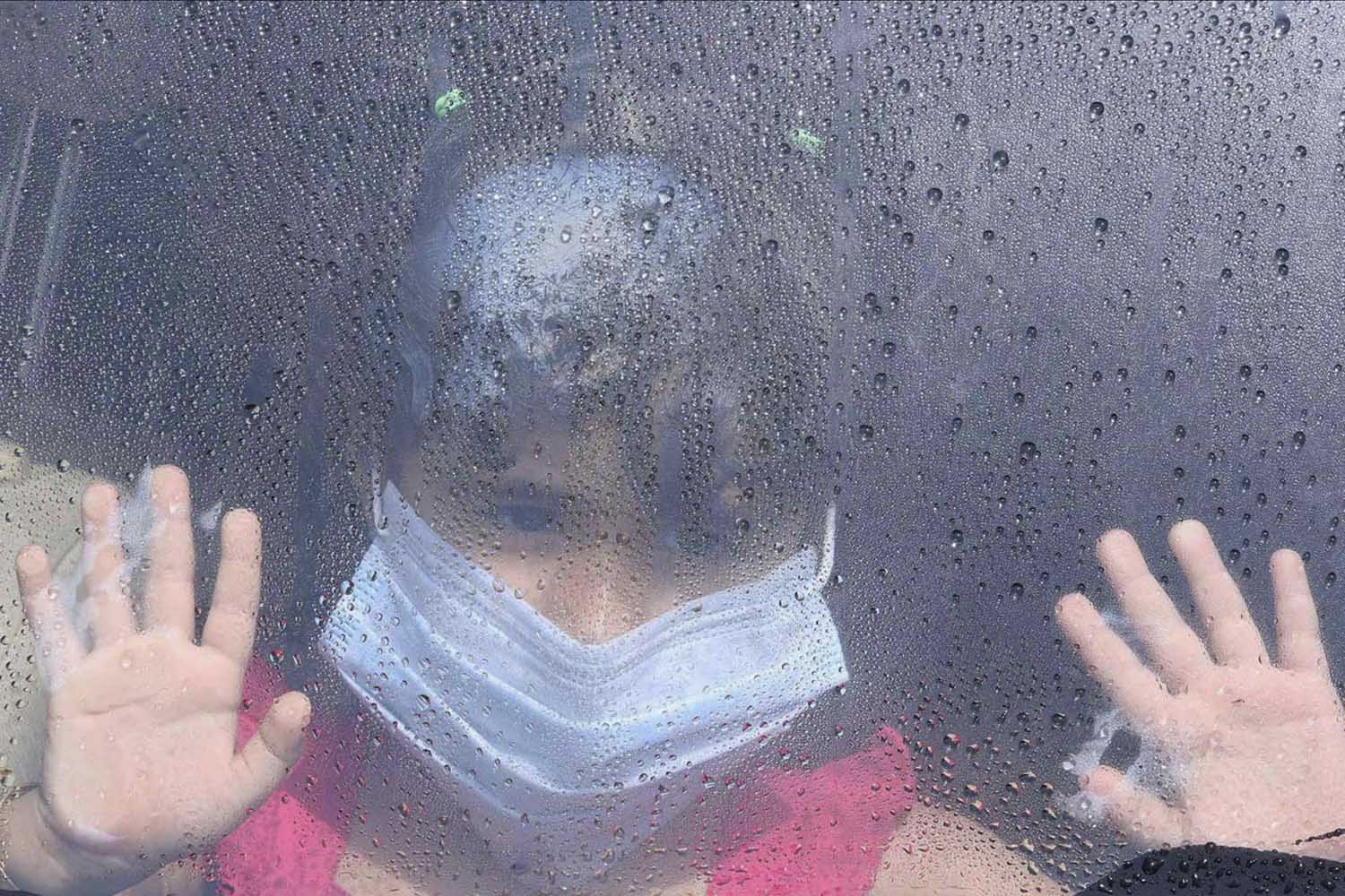 A girl wearing a mask looks out a car window as rain drops are seen in Taif