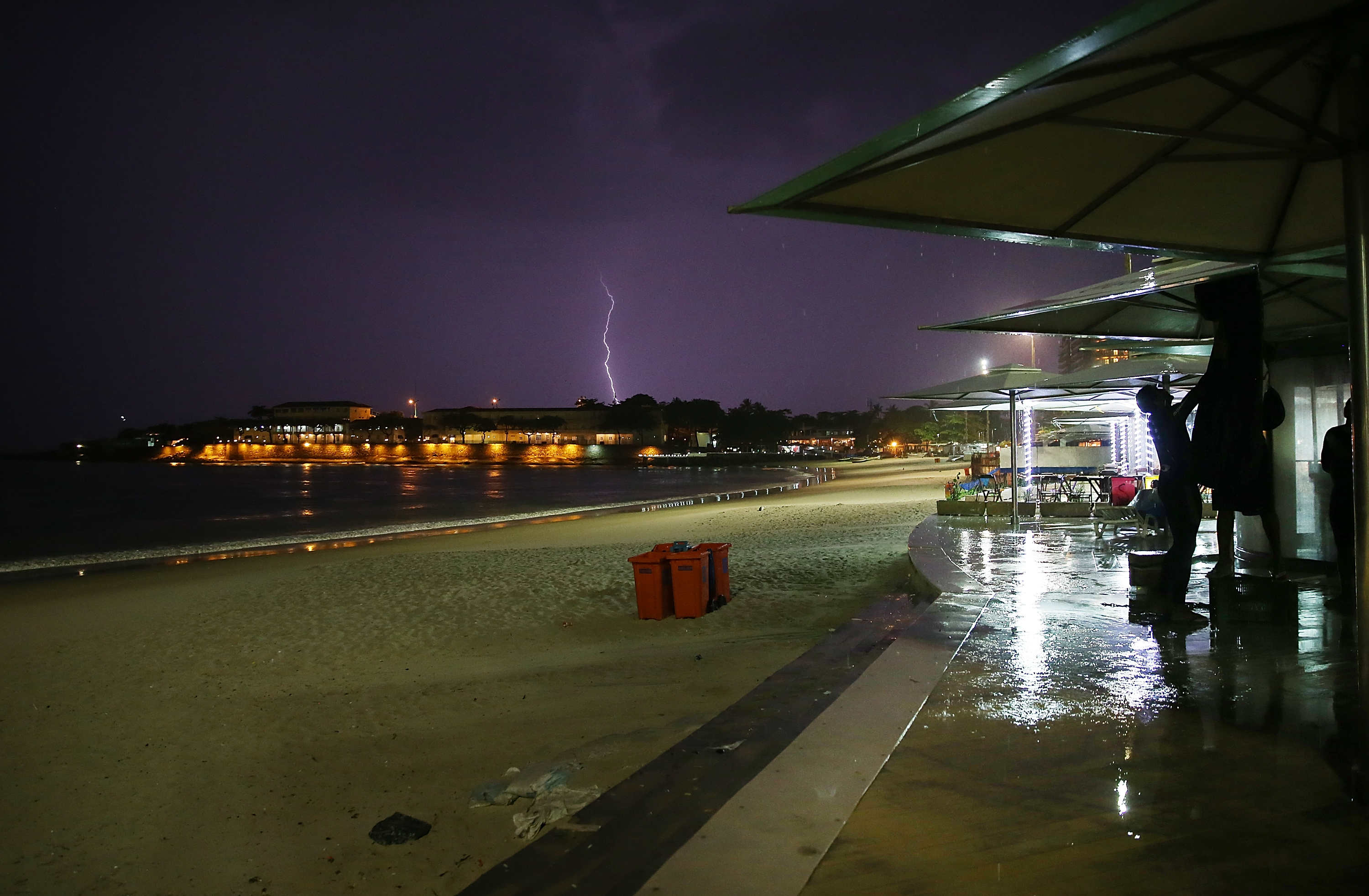 A worker stands beneath umbrellas along Copacabana Beach as lightning flashes during a powerful early evening thunderstorm on January 16, 2014 in Rio de Janeiro.