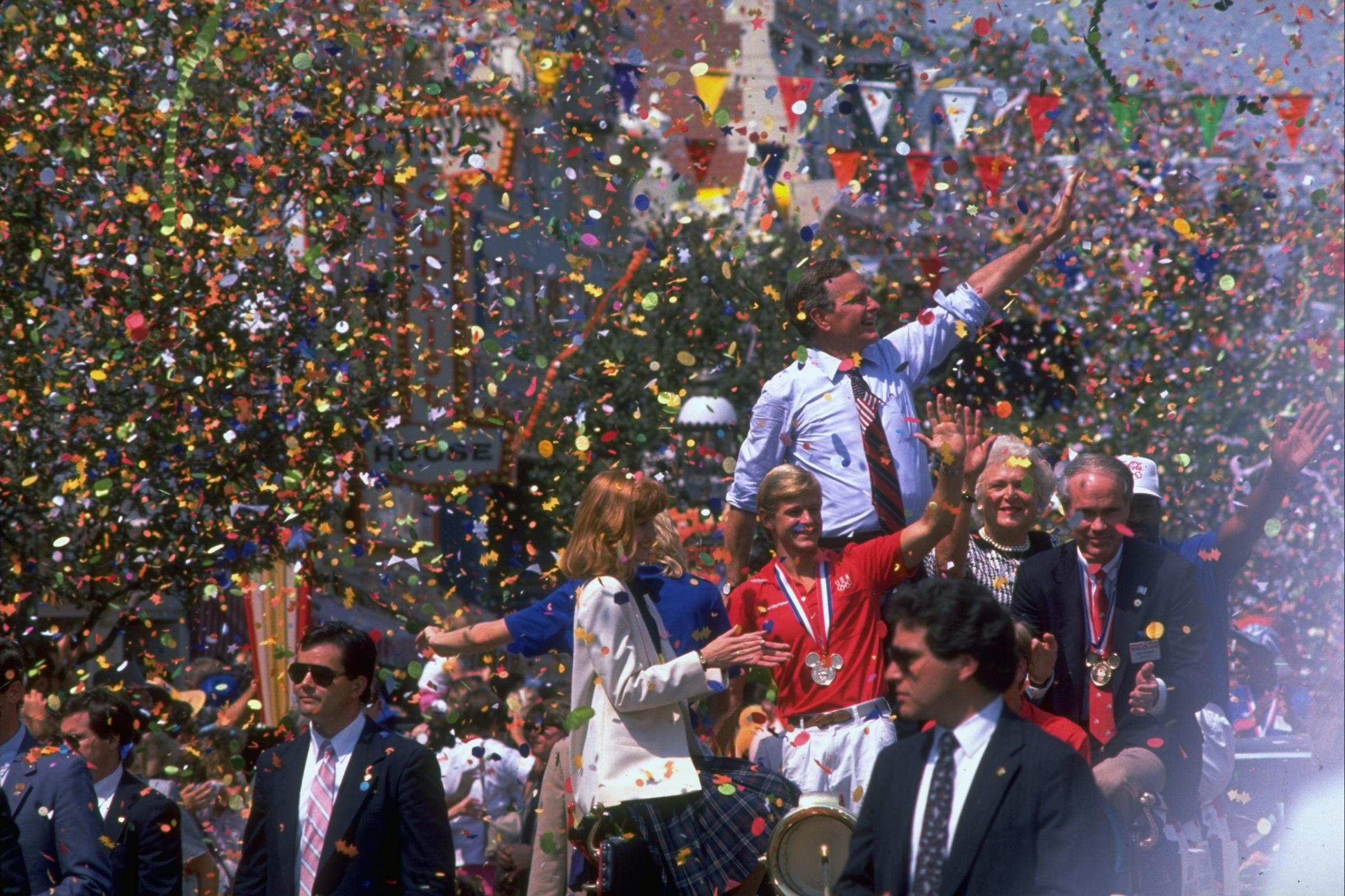 Republican presidential candidate George H.W. Bush waving above a crowd amid shower of parade confetti, with wife Barbara by his side at a send-off gala for the US Olympic team.