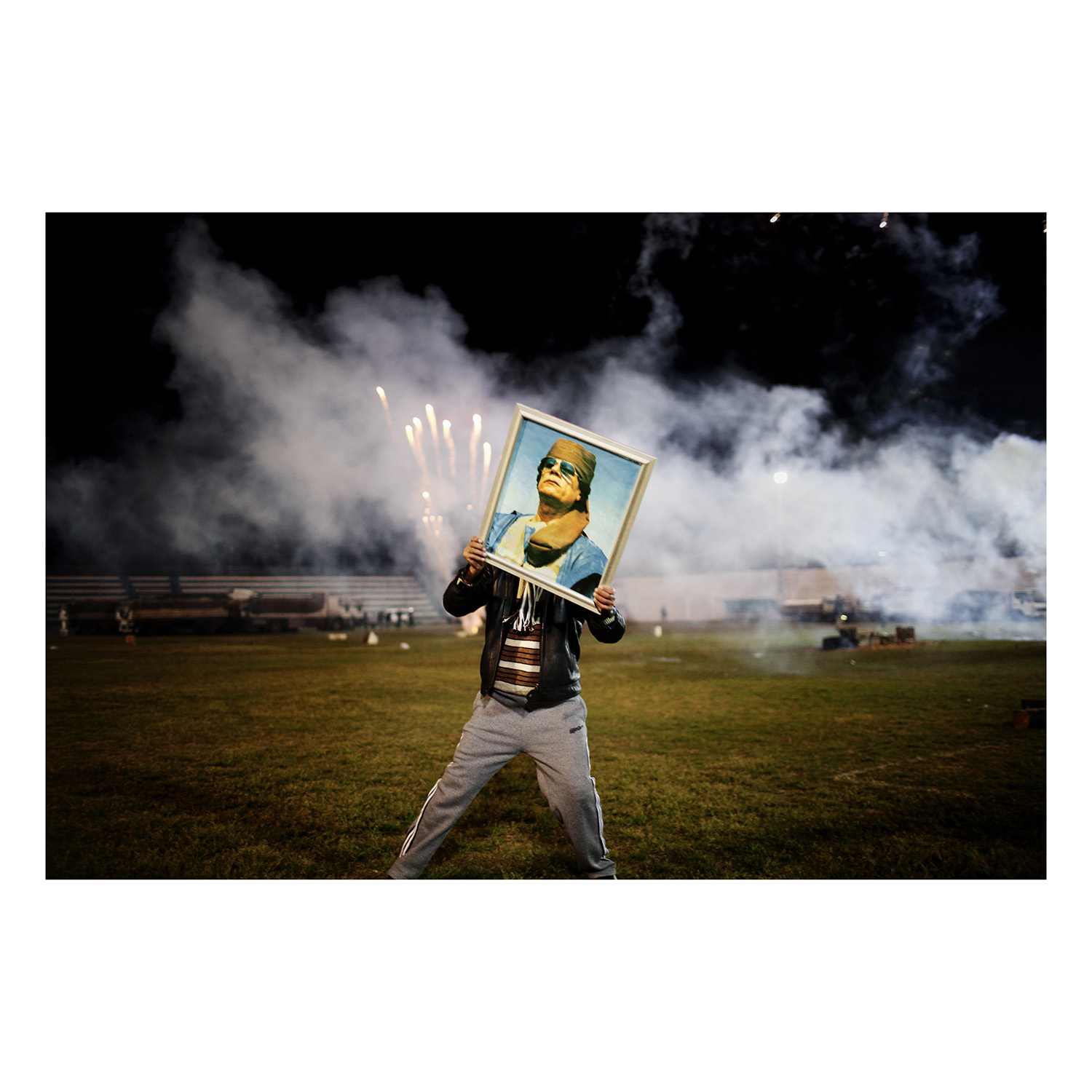 LIBYA. March 9, 2011. A Qaddafi supporter holds a portrait of the Libyan leader as fireworks go up in the background on a soccer field in a suburb of Zawiyah where government minders took a group of foreign journalists to attend a staged celebration.