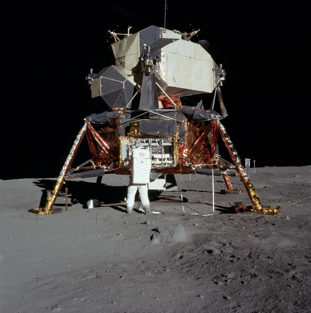 Buzz is preparing to remove the seismometer from the lefthand compartment in the equipment bay. He used a pulley-mounted tape to raise the door which can be seen above the right side of the bay. The tape is visible to his right, draped over a LM strut.