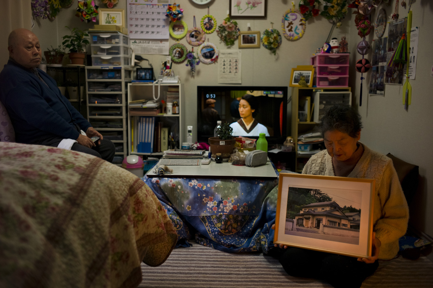 Japan, Aizuwakamatsu, 2014. Idogawa Ikuko 82 (right) holds a picture of their old house which was located in Okuma and now too radiated to return to as her husband Idogawa tsuguo 85 (left) sits with her in their sleeping and living room decorated with trinkets and hand made crafts in the temporary housing they were assigned to.