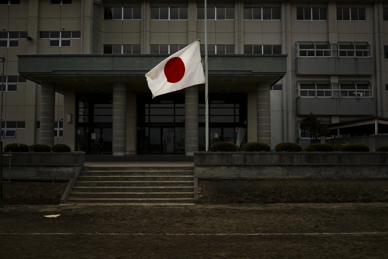 Japan, Minamisoma, 2014.
                              A Japanese flag flies half-mast at a school on the third anniversary of the earthquake and tsunami that hit the northeastern coast of Japan on March 11th 2011. The school is approximately 30 kilometers from the damaged Fukushima Daiichi Nuclear Power Station. Many original residents have returned to their homes despite the  high levels of radiation recorded in the area and the effect that exposure to it may have on their health.