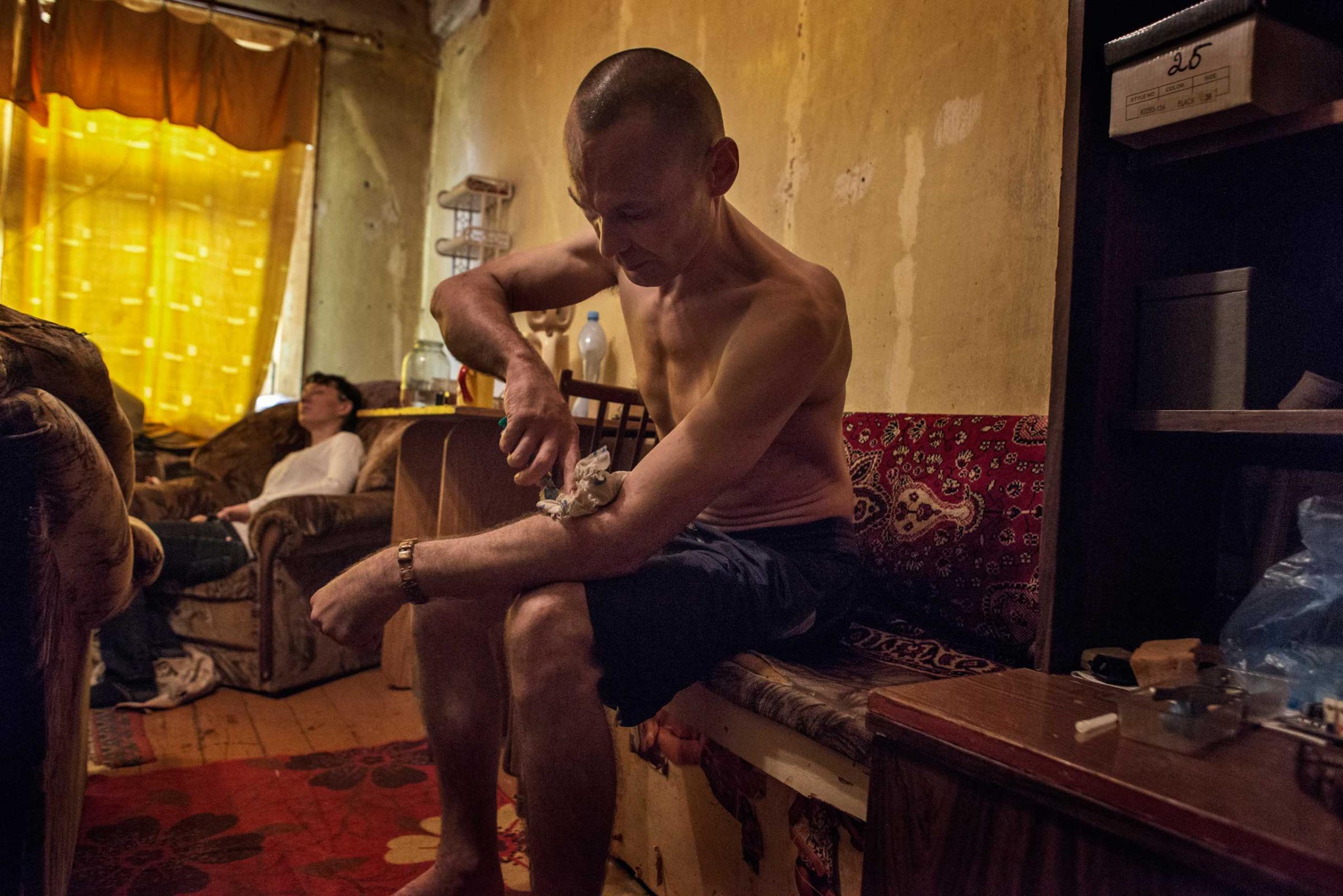 Andrey injects krokodil, with Zhanna in the background already under the influence of the drug.