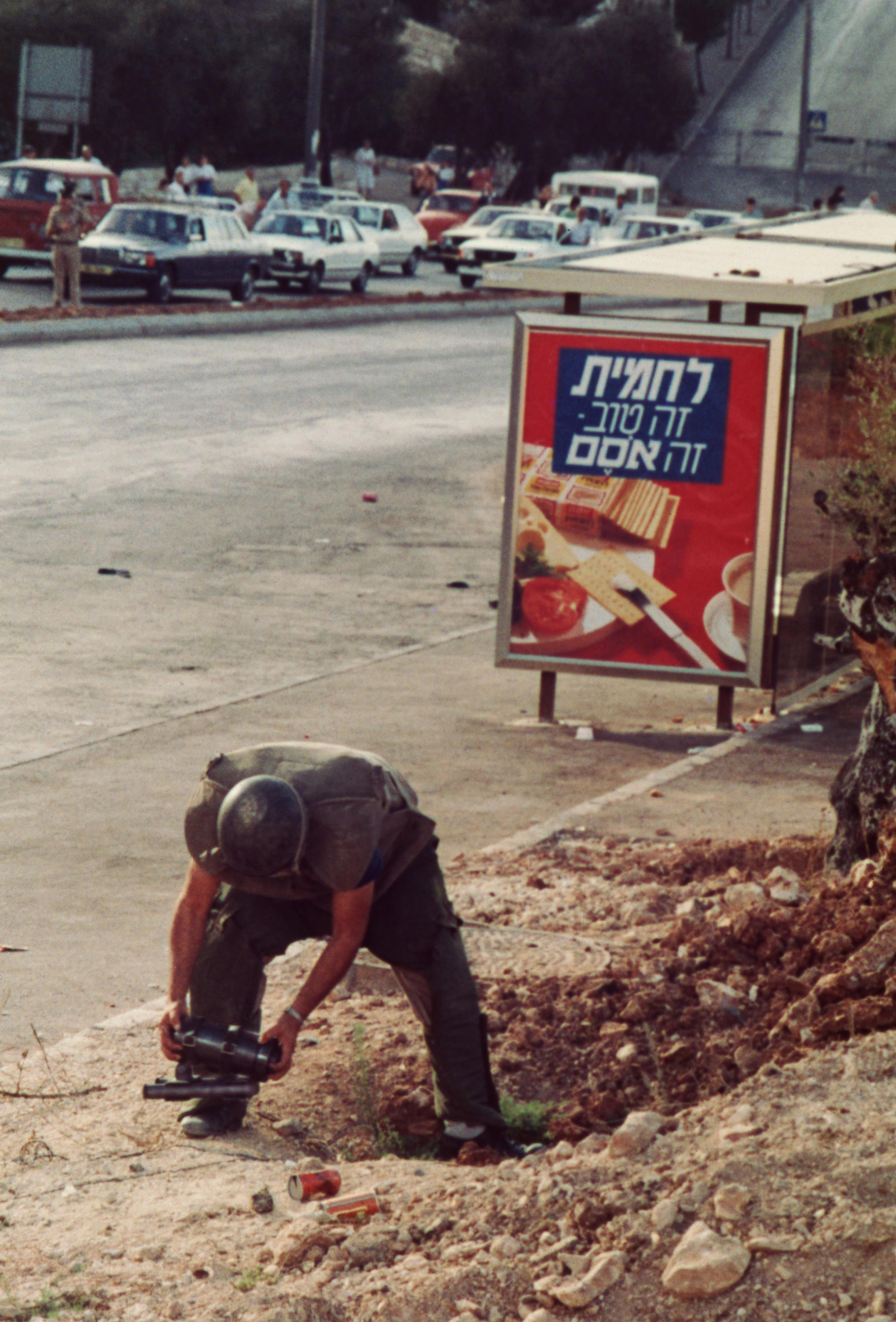 Traffic in downtown Jerusalem comes to a halt beside a bus stop while one disposal expert named Uri, 32, gingerly handles the apparent bomb, 1984.