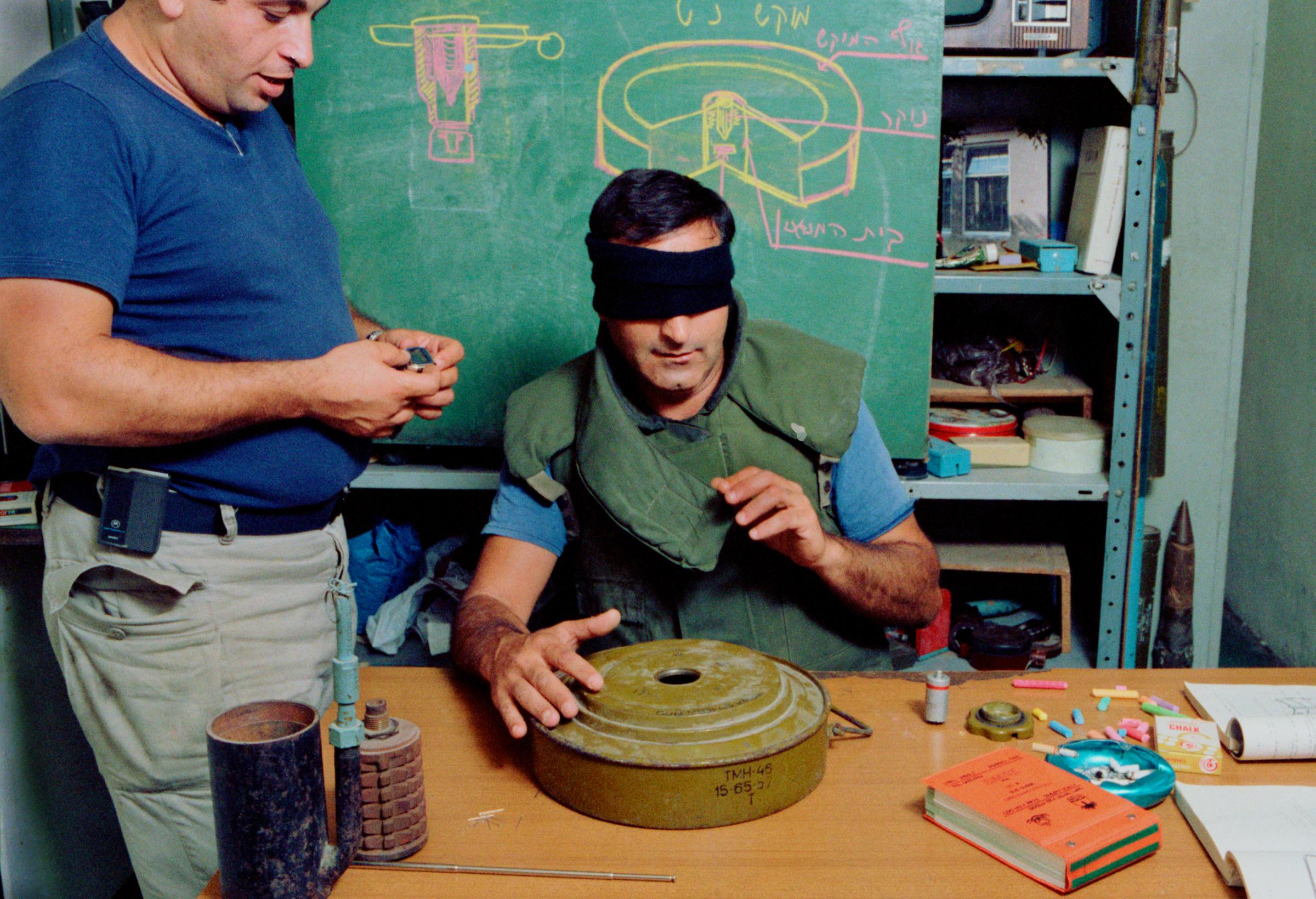 Commander Amedi oversees the test of one bomb squad member who must be able to recognize and disarm a wide variety of bombs by his sense of touch alone. The first example on his table, already successfully dismantled, was a Soviet-style Bouncing Betty, while the one that currently has his full attention is an Israeli-made IM-46 land mine big enough to blow up a truck.