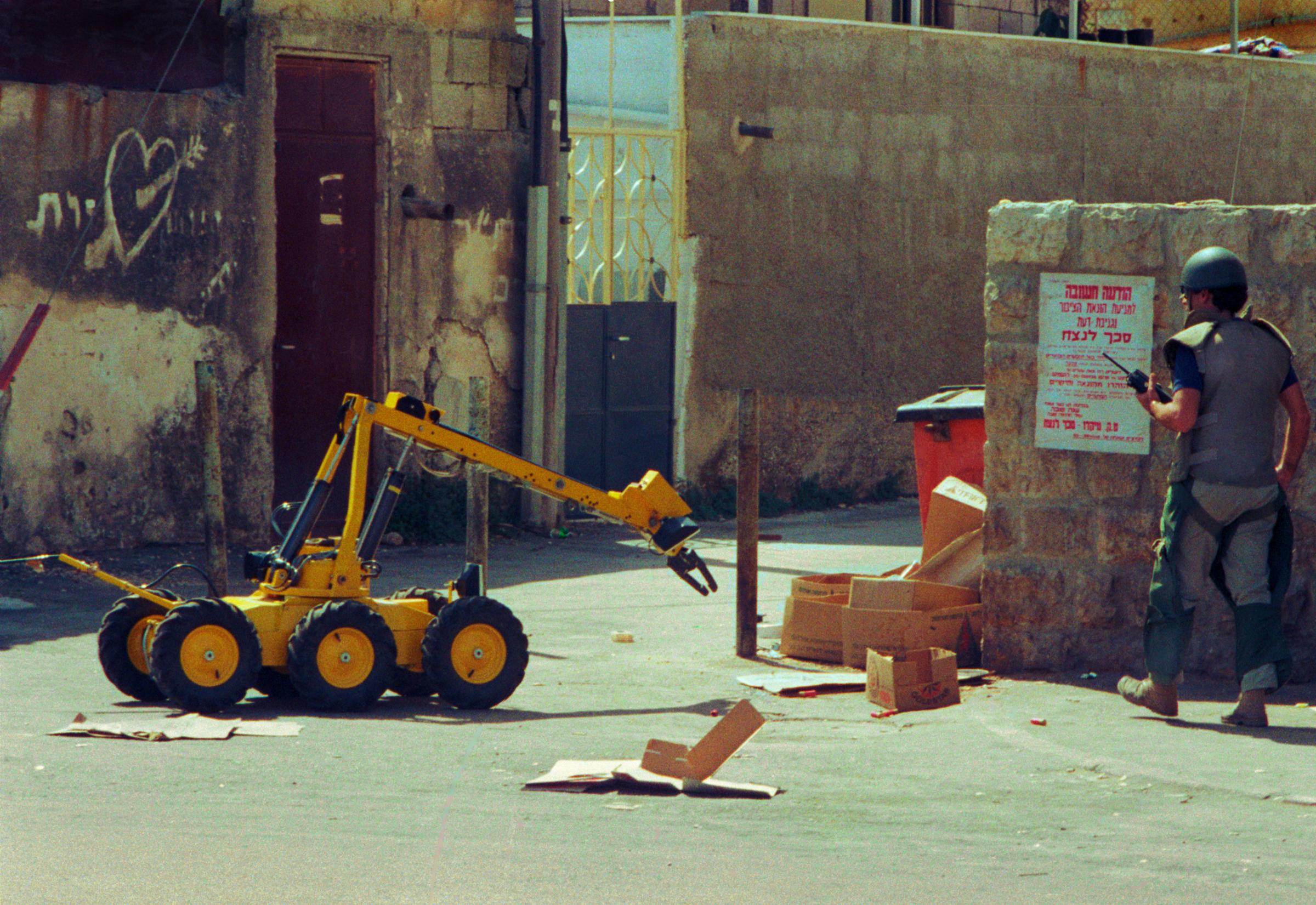Uri watches as a shotgun-toting robot is aimed, with the aid of a remote video camera, at a possible bomb near the trash can in a Jerusalem side street.