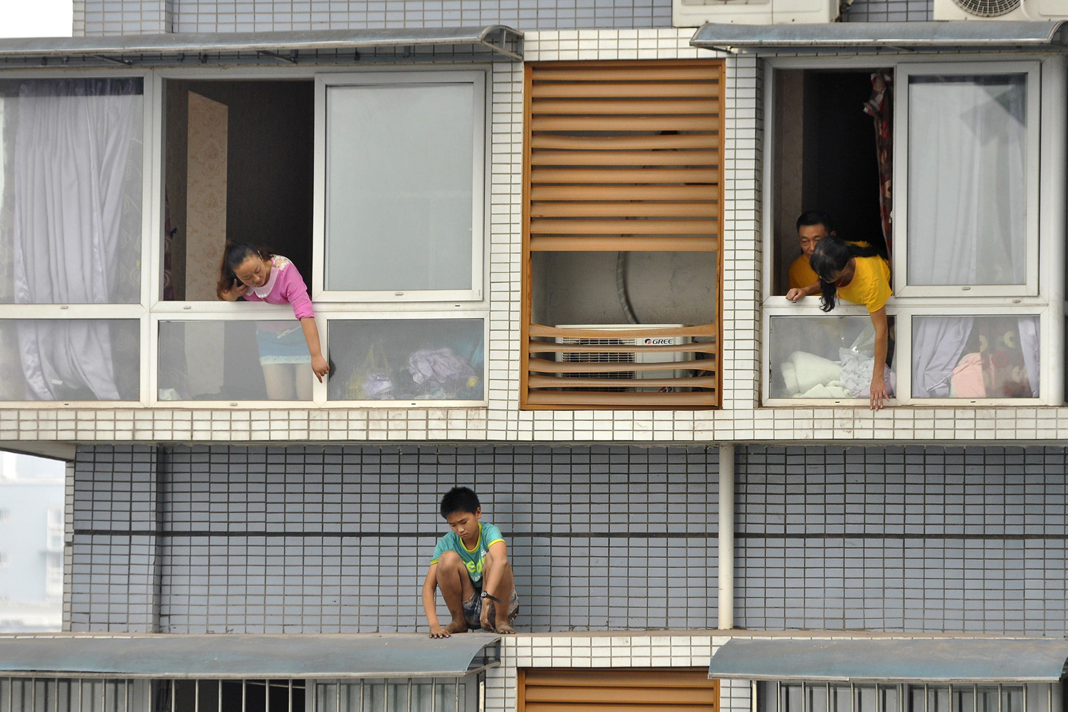 A twelve-year-old boy sits outside a window of his eleventh-floor apartment as his relatives try to ask him to come back back inside, in Yibin