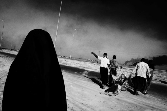 Paolo Pellegrin, March 4, 2003I entered Iraq unembedded with a car I had rented in Kuwait, stopping to photograph the fighting in Basra, a city in the South on the highway to Baghdad. The picture was taken near some sort of compound where there had been fighting between pro-Saddam fighters and British forces. There were several bodies of Iraqi fighters lying around. At one point, people started to appear on the streets to drag away particular bodies. As I understand, the woman in the foreground of the photograph was the mother of the deceased. They dragged him from the place he was killed, put him in the trunk of a waiting car, and drove off. When I look at this image ten years later, the first thing that comes to mind is the idea of loss. I see the photograph and think of the mother's loss. If I continue looking, that black veiled figure, in some strange sense, makes me think of death itself Ñ shadowy and dark.