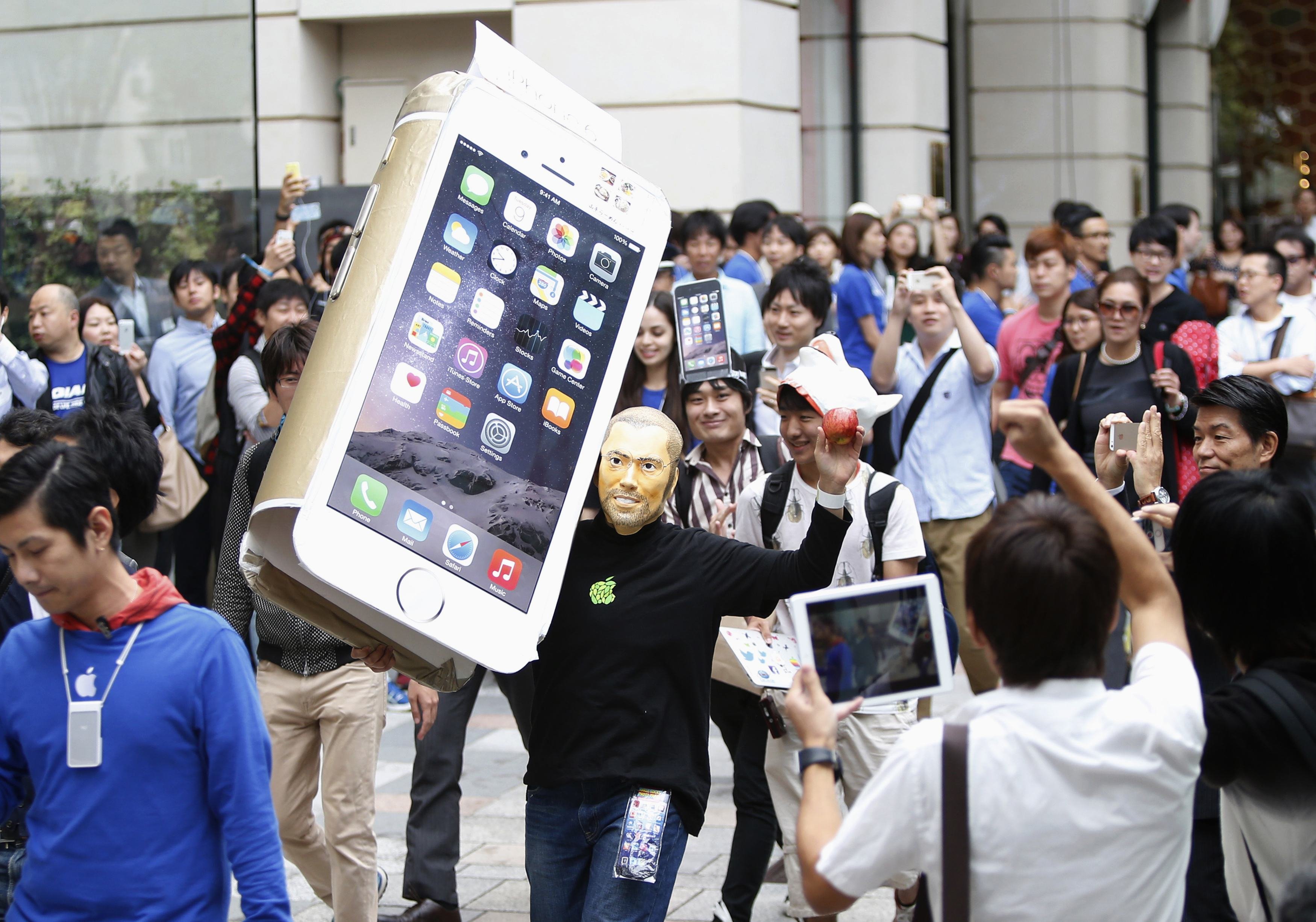 A man wearing a mask depicting Apple's co-founder Steve Jobs holds up a cardboard cut-out of Apple's new iPhone 6, as he walks into an Apple Store in Tokyo on Sept. 18, 2014.