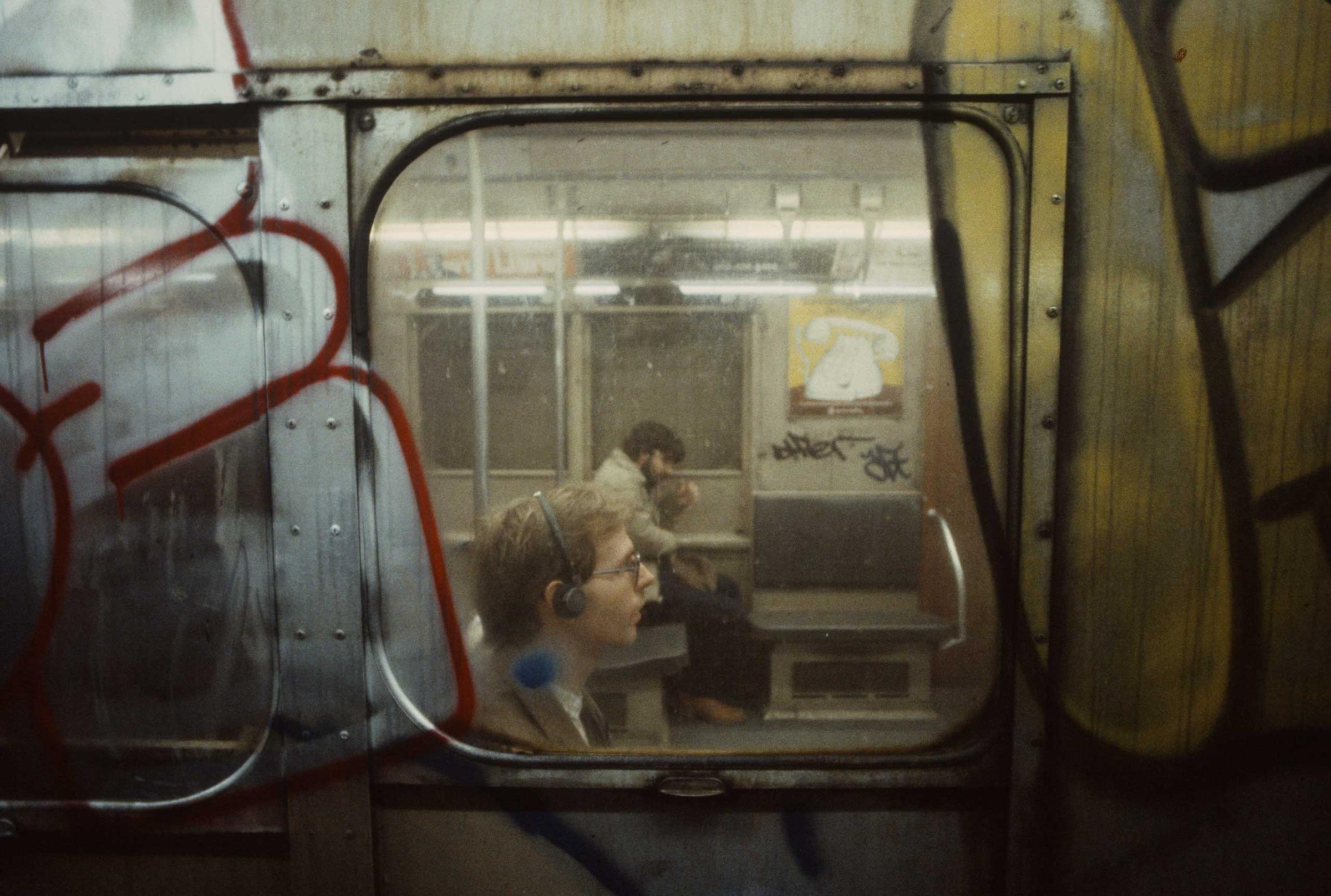 A man is seen wearing headphones through a subway car window, 1981.