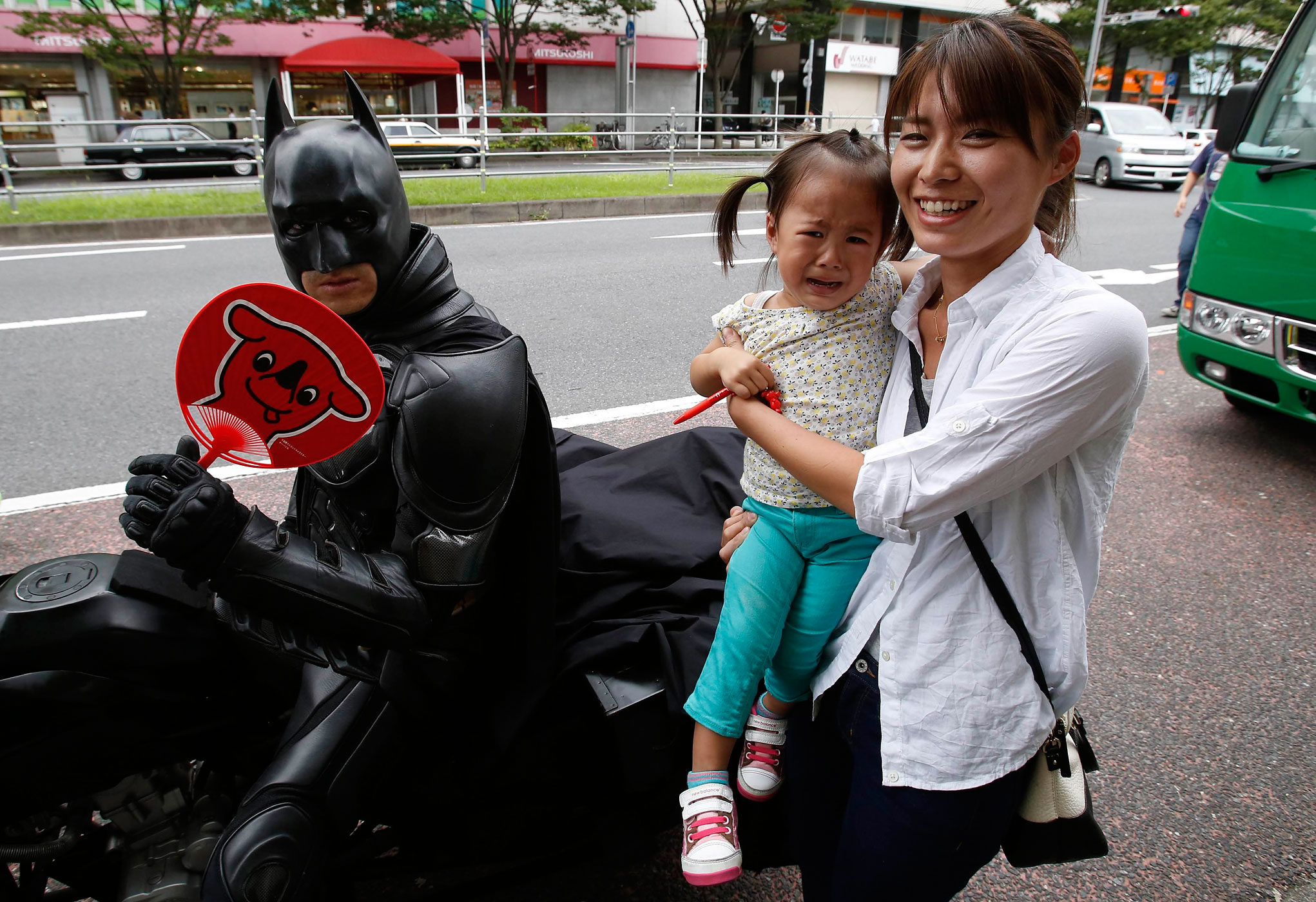 A woman carrying a crying child poses for pictures next to Chibatman.