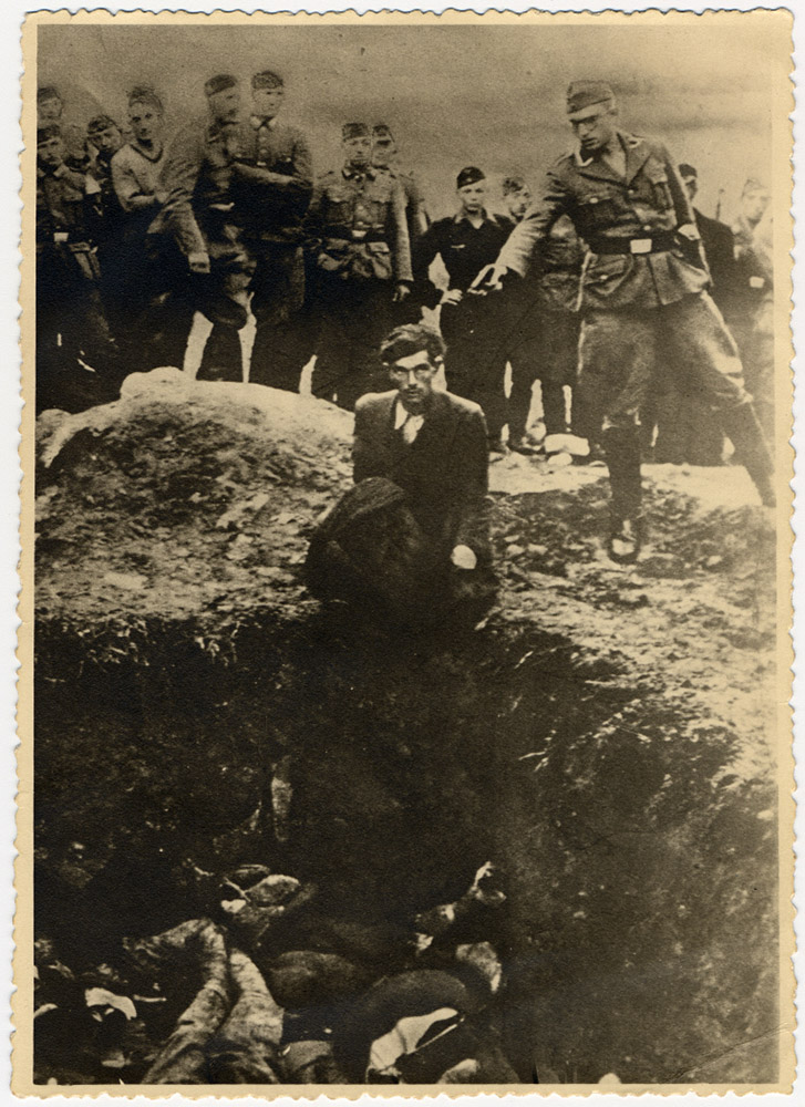 German soldiers of the Waffen-SS and the Reich Labor Service look on as a member of an Einsatzgruppe prepares to shoot a Ukrainian Jew kneeling on the edge of a mass grave filled with corpses, Vinnitsa, Ukraine, c. 1941-43.