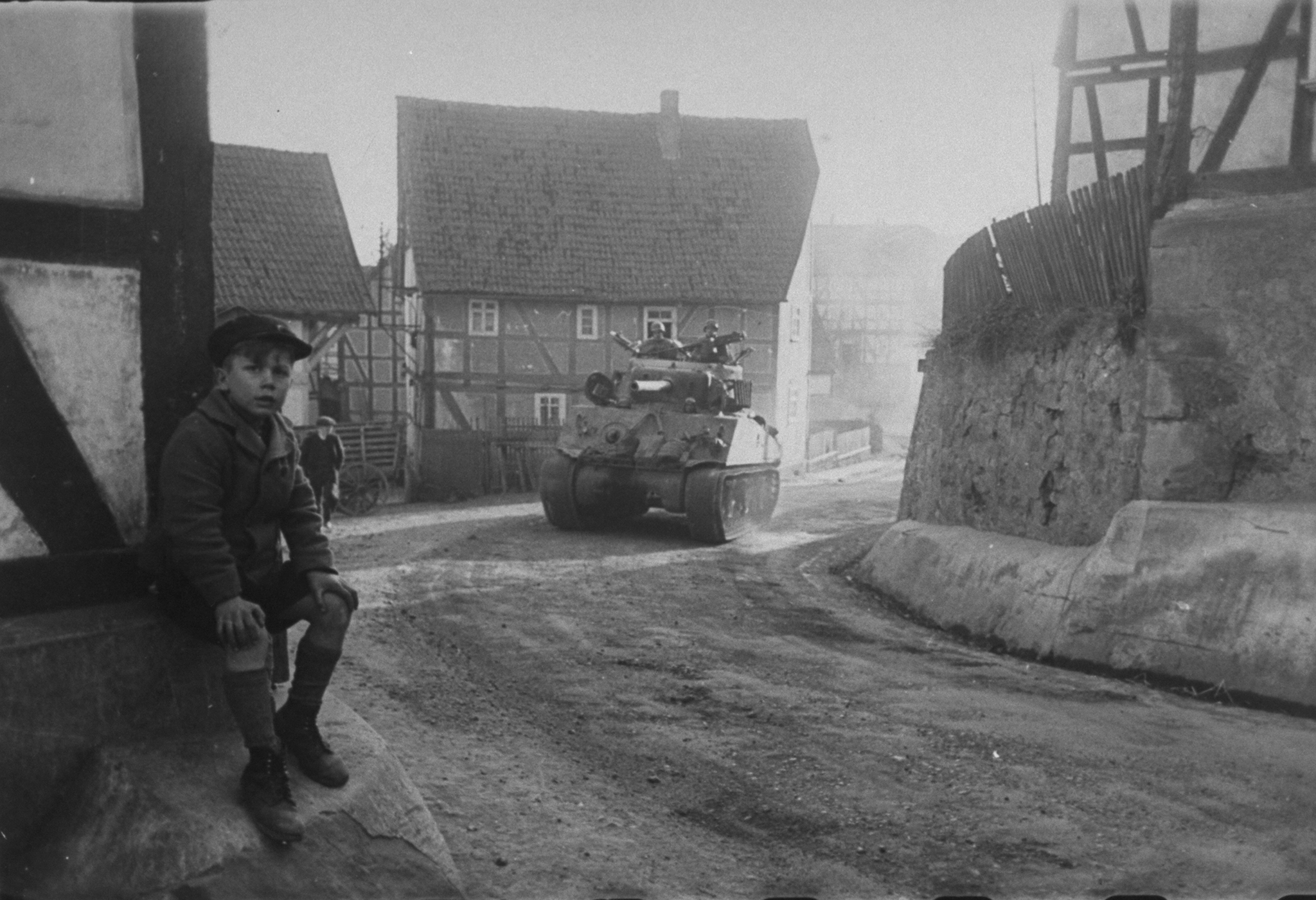 A young German boy sits beside the road as a tank of the U.S. 9th Armored Division passes through his village on its way to Berlin, spring 1945.