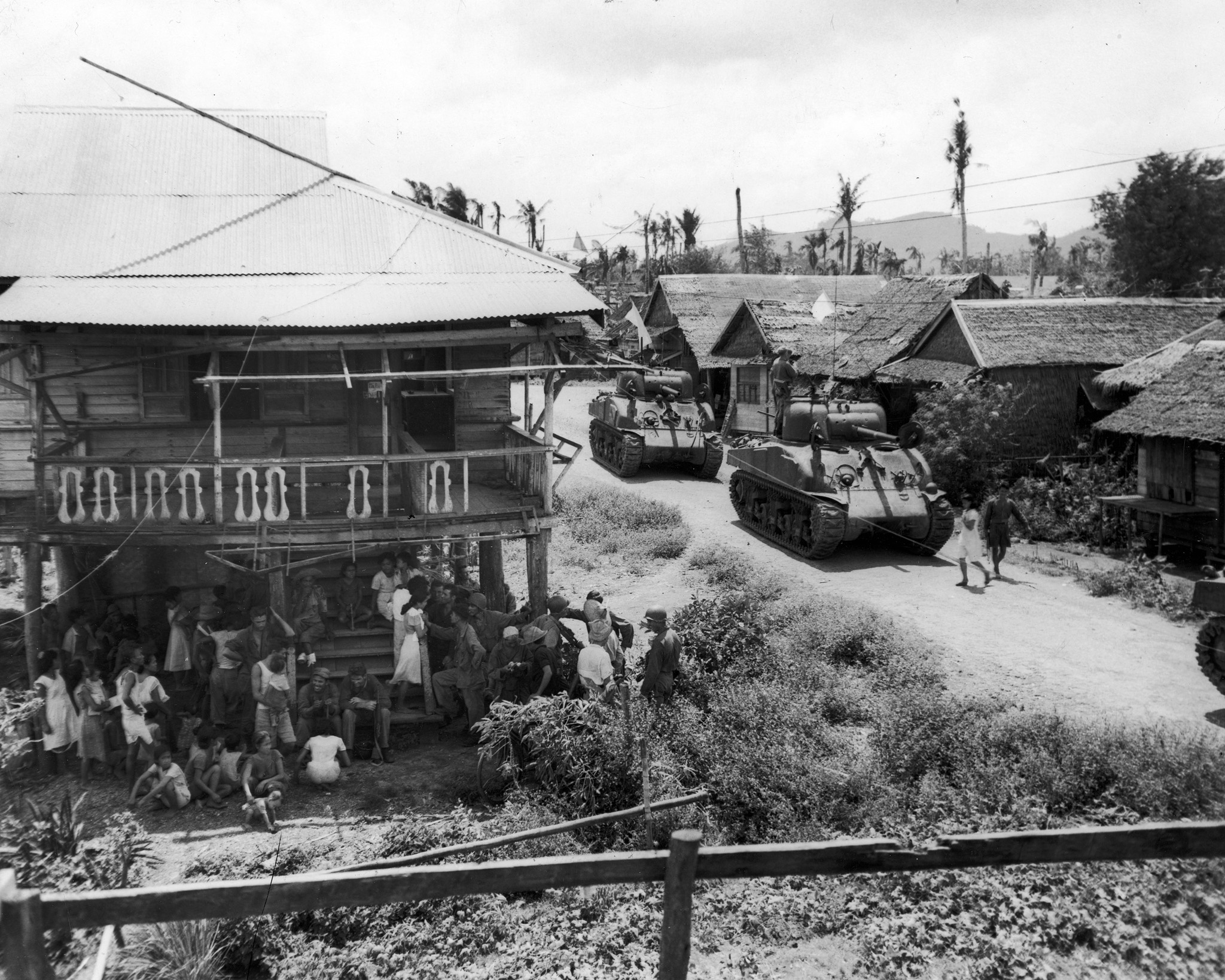 Villagers chat with American soldiers after being freed from Japanese occupation while Sherman tanks of the 1st Cavalry Division follow the retreating enemy, Philippines, October 1944.