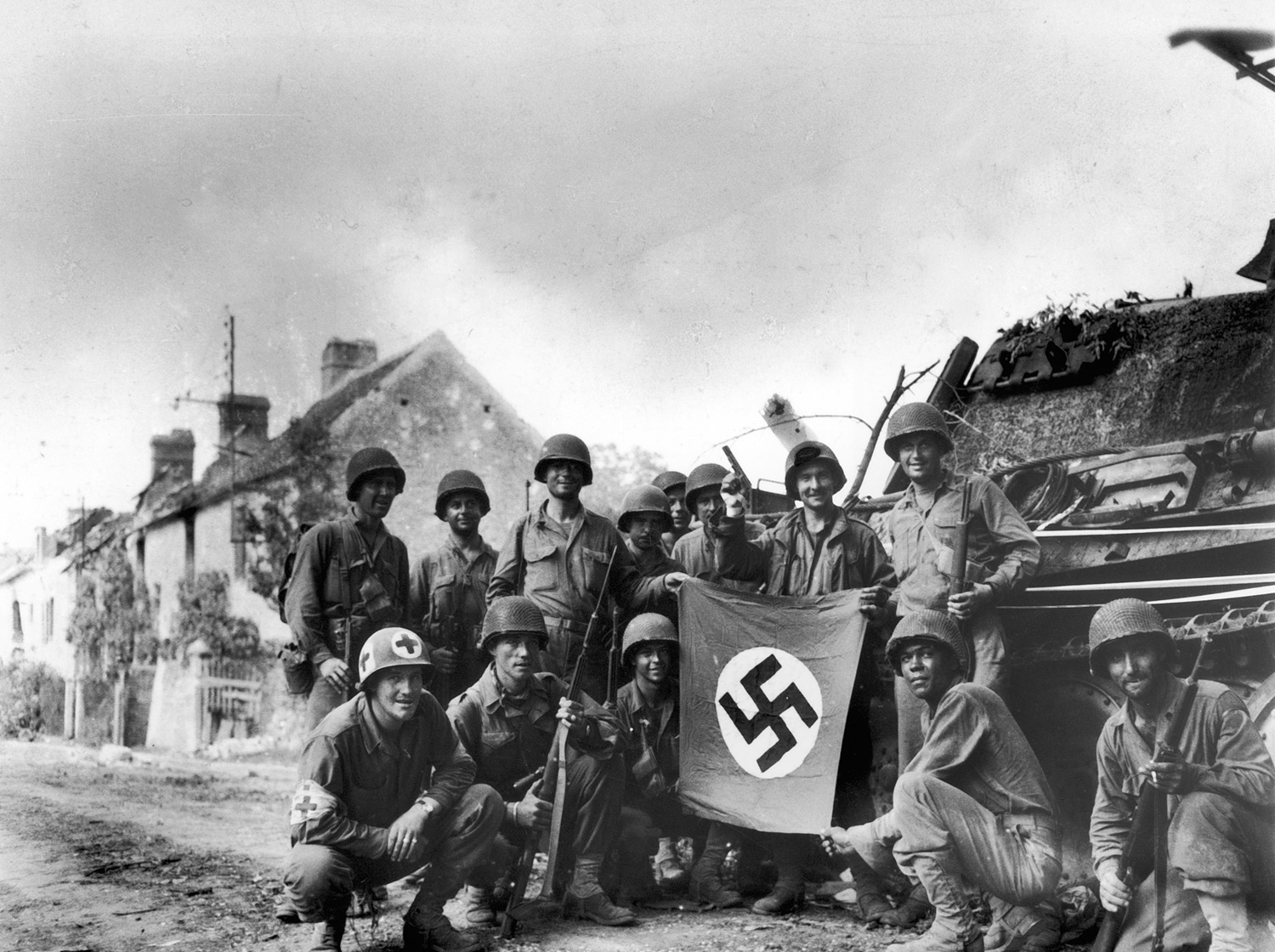 American infantrymen, left behind to mop up last stronghold of Nazis in Falaise Gap area of northwestern France, line up in front of wrecked German tank with a captured swastika flag, 1944.