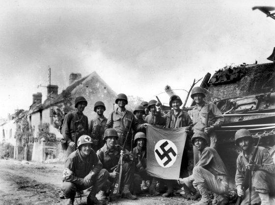 American infantrymen, left behind to mop up last stronghold of Nazis in Falaise Gap area of northwestern France, line up in front of wrecked German tank with a captured swastika flag, 1944.