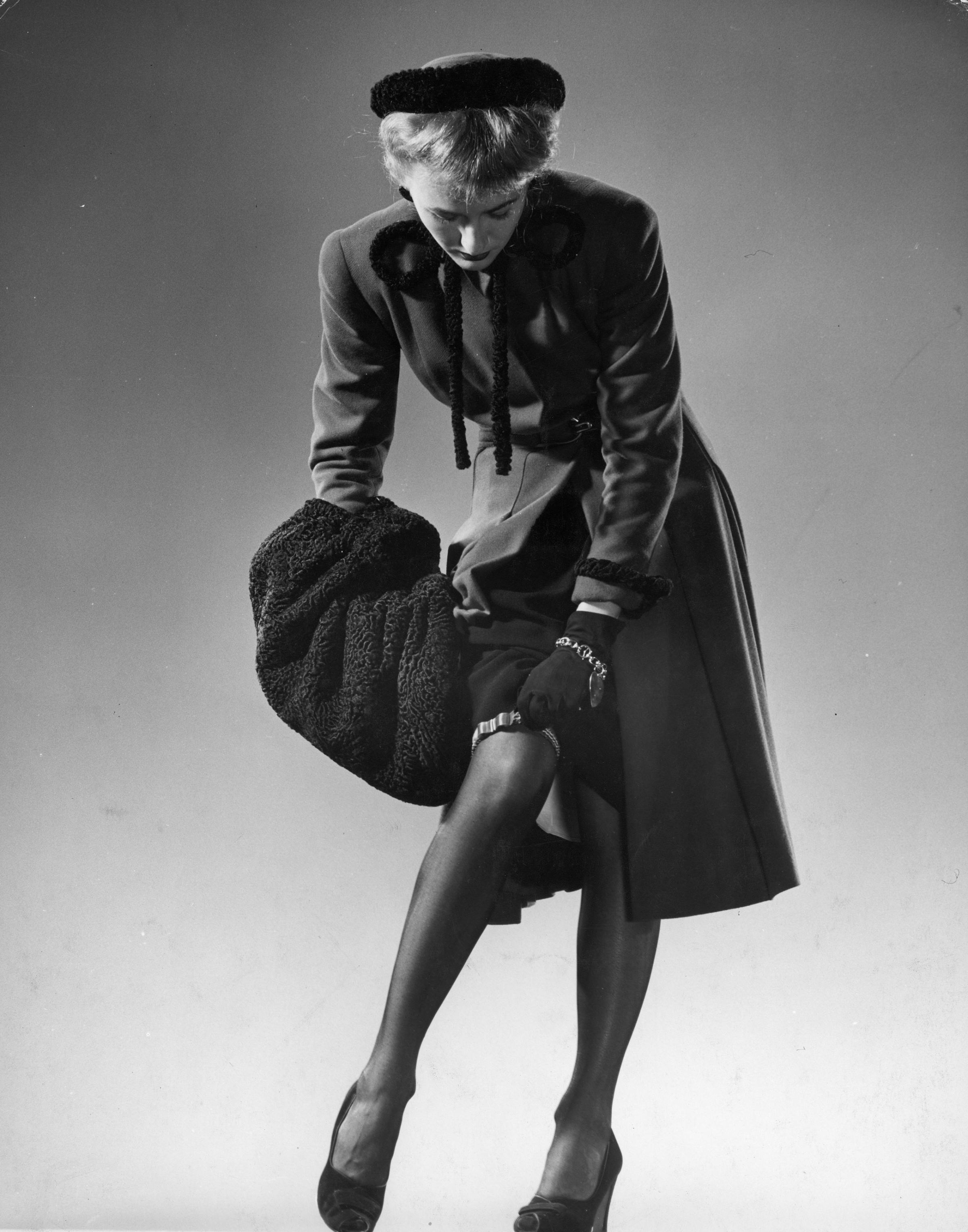 Model wearing black stockings, fitted dress coat, fur trimmed hat and fur muff, adjusting garter, 1946.