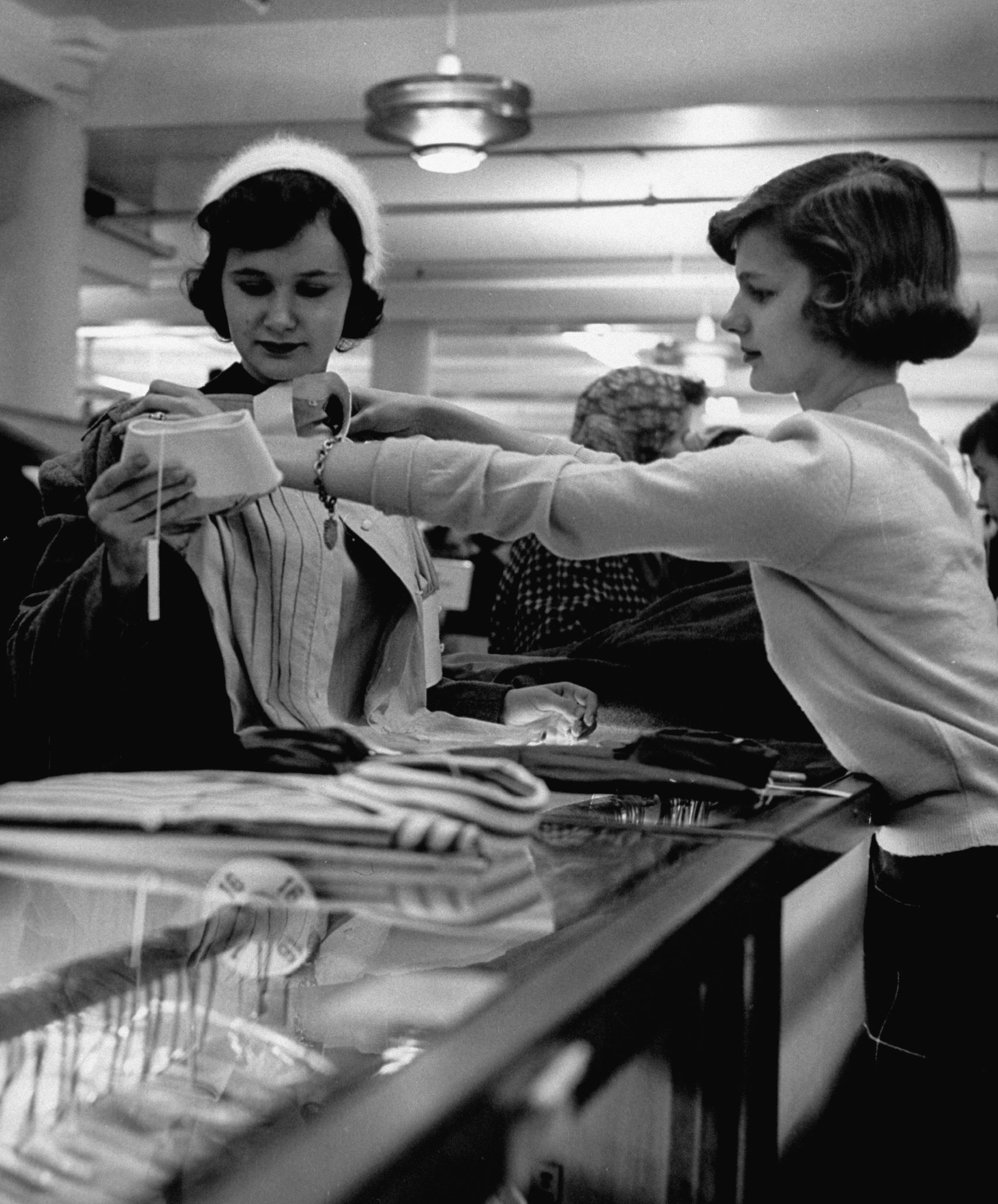 Attractive young sales girl holding blouse up to customer in store, as customer is looking at other things to buy.