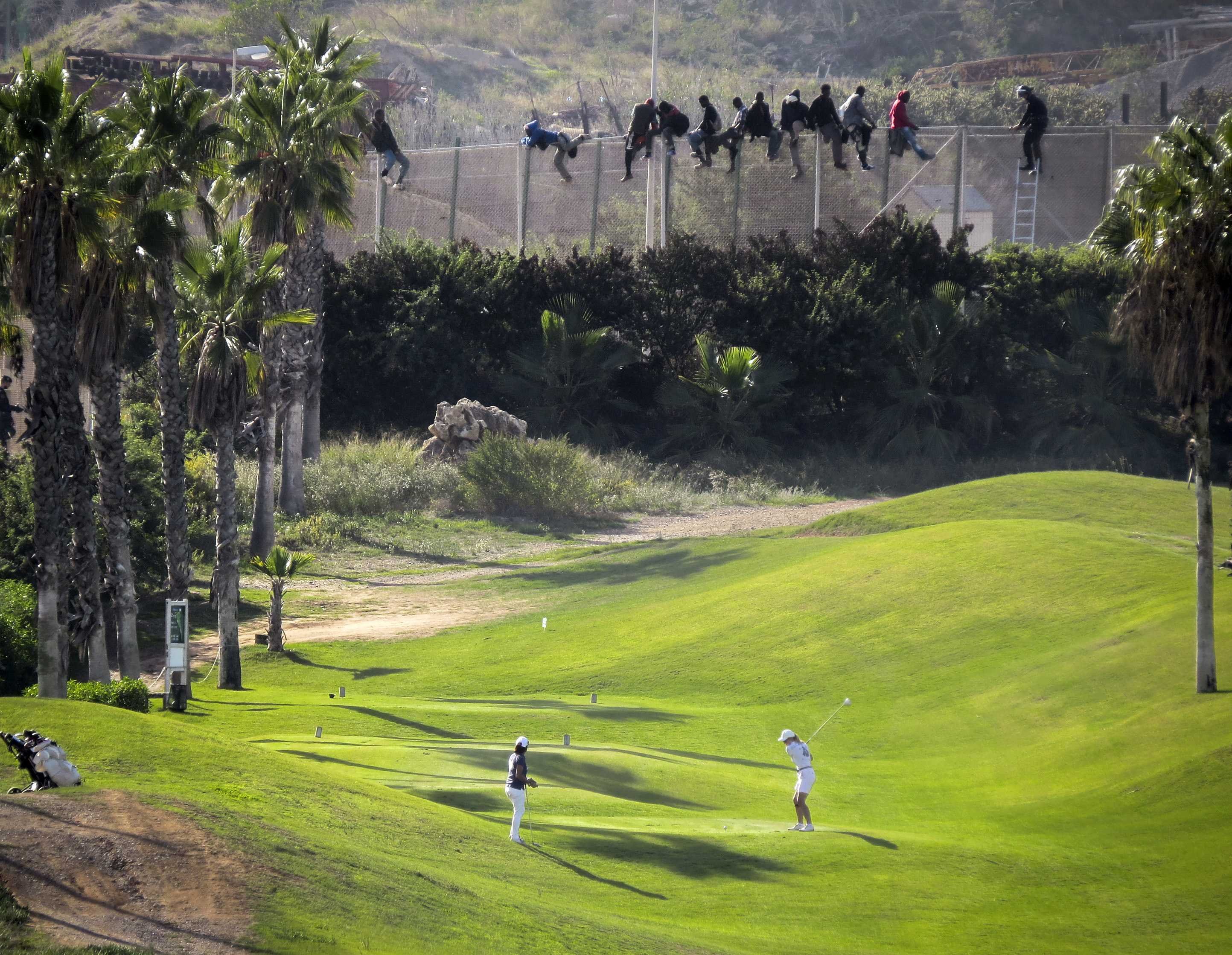 A golfer swings as African migrants sit atop a fence during an attempt to cross from Morocco into the Spanish enclave of Melilla on Oct. 22, 2014. (José Palazón—Reuters)