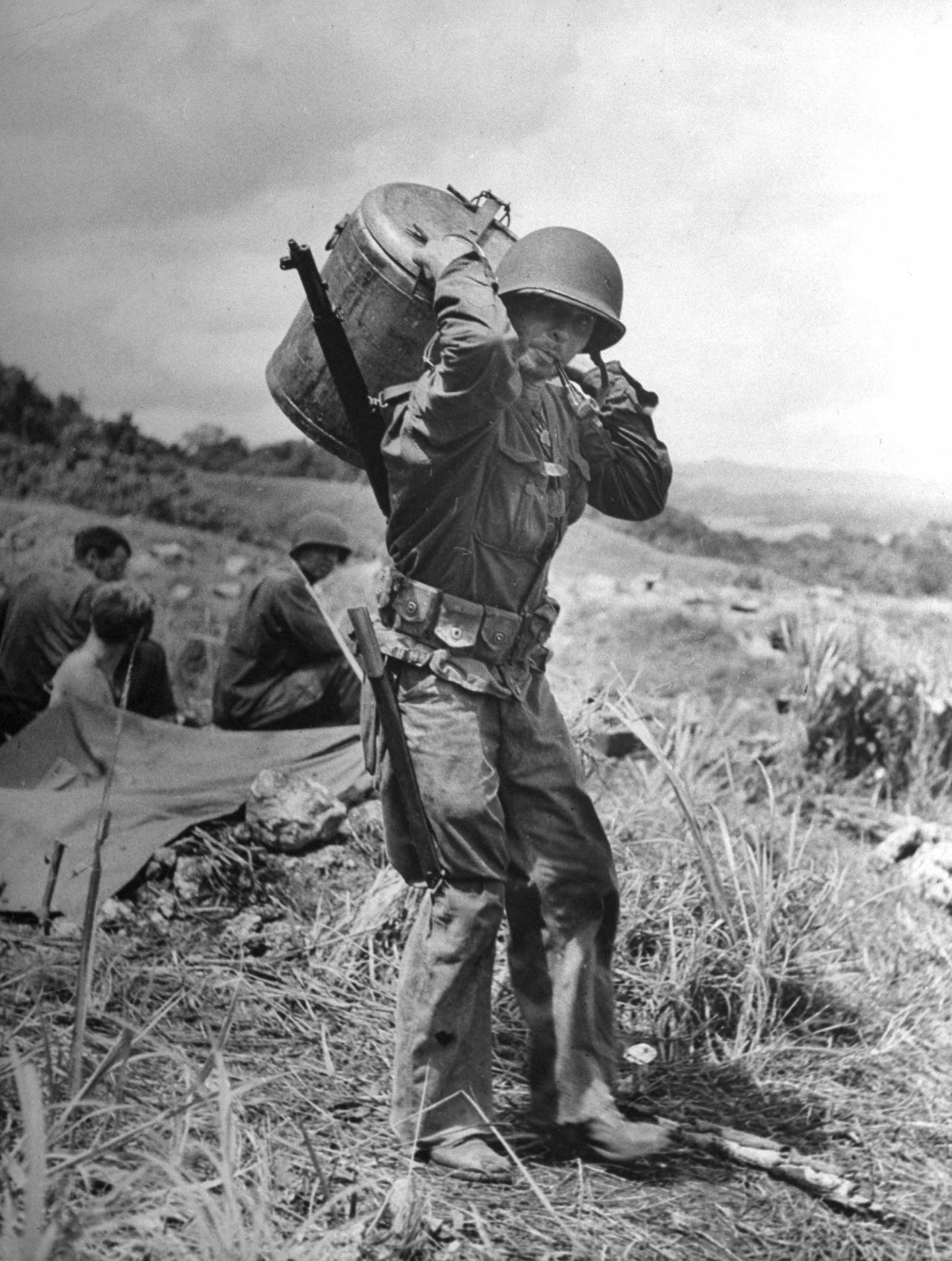 "On his back a wiry, stooped U.S. infantryman, smoking an old pipe, carries a heavy container filled with supplies up over the hills to the front."
