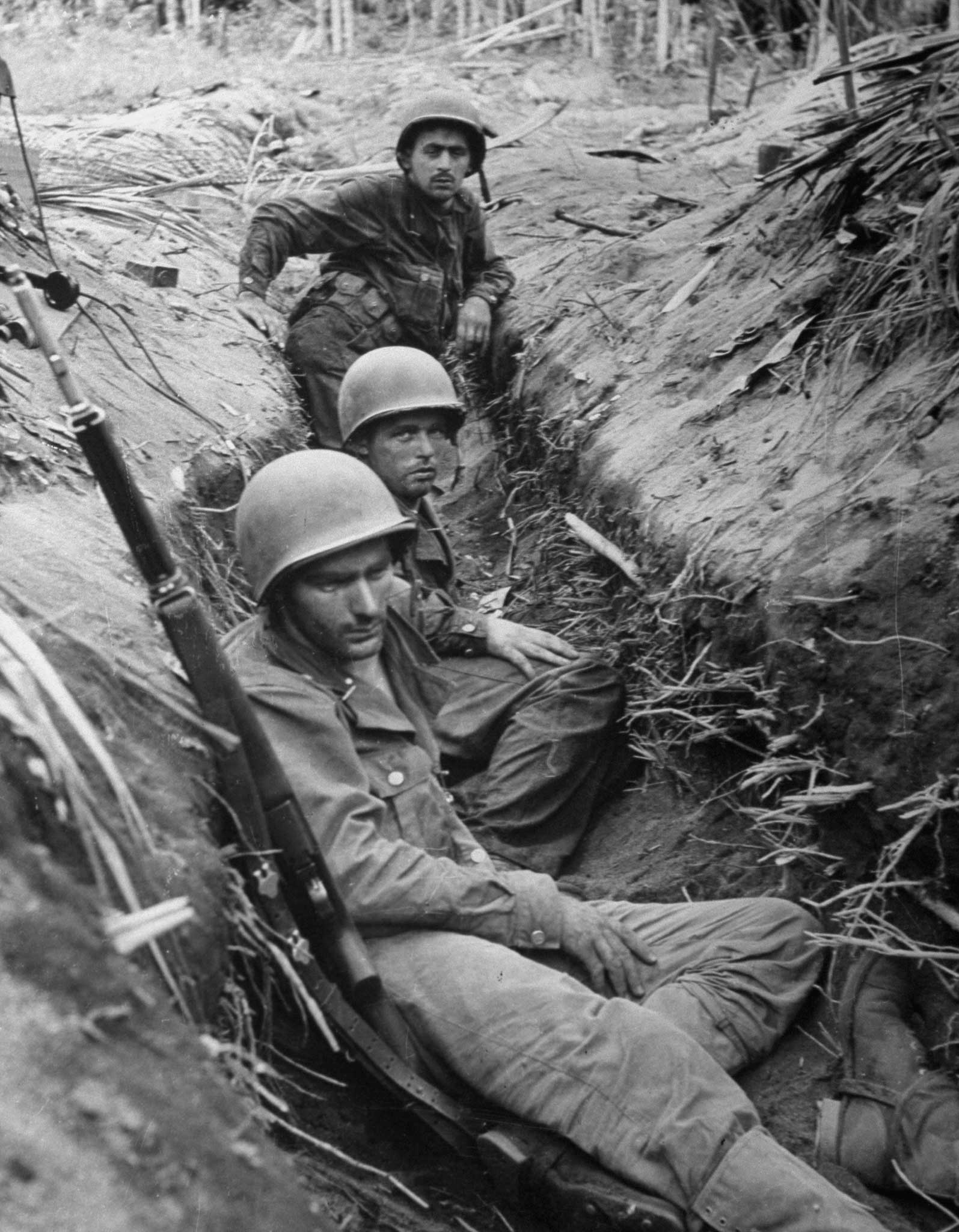 "In a captured Jap intercommunication trench, soldiers rest and clean their guns. Sniping is going on right above them."