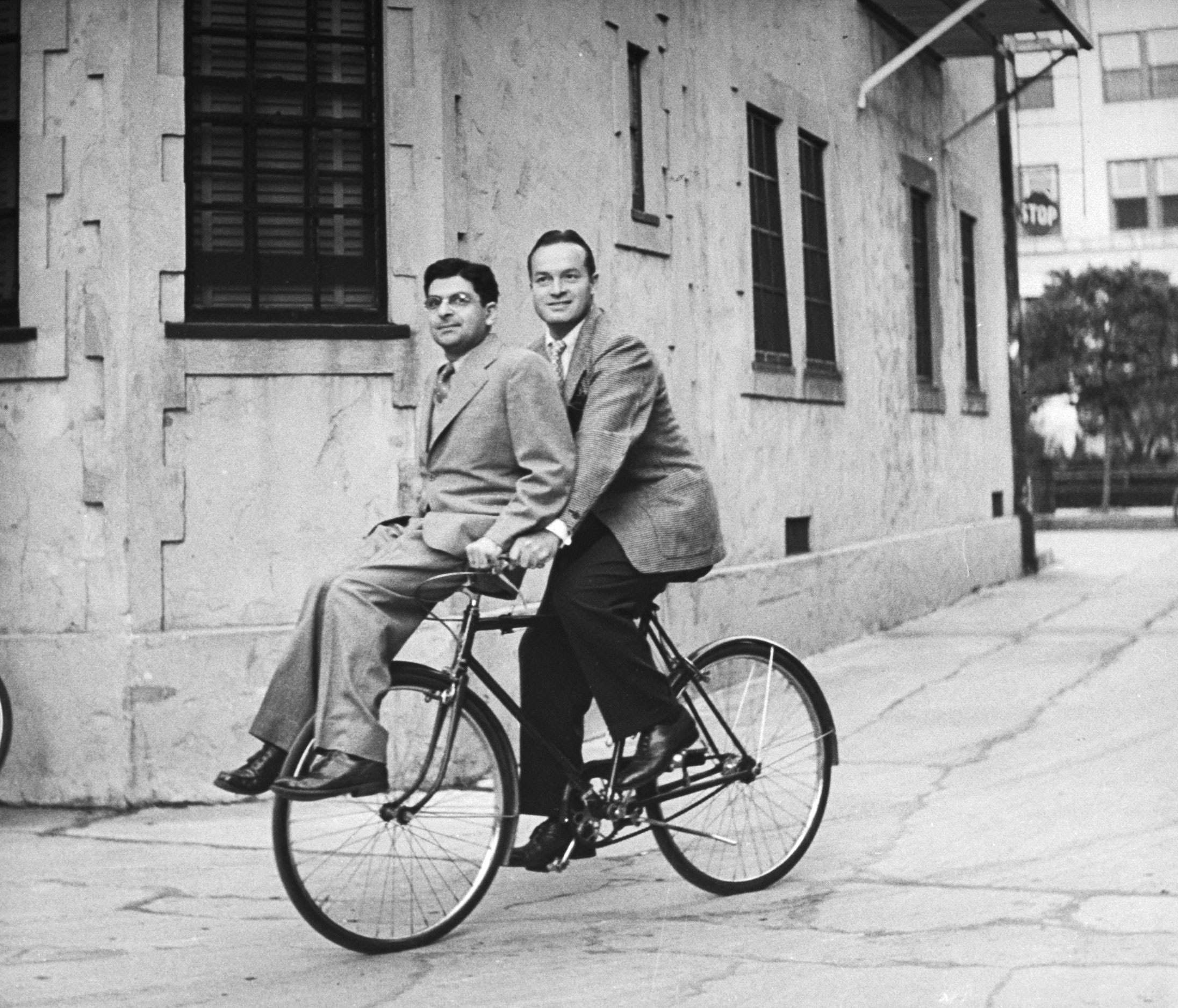 Columnist Sidney Skolsky catching a ride on Bob Hope's bicycle on Hope's way from his dressing room to a sound stage, 1943.