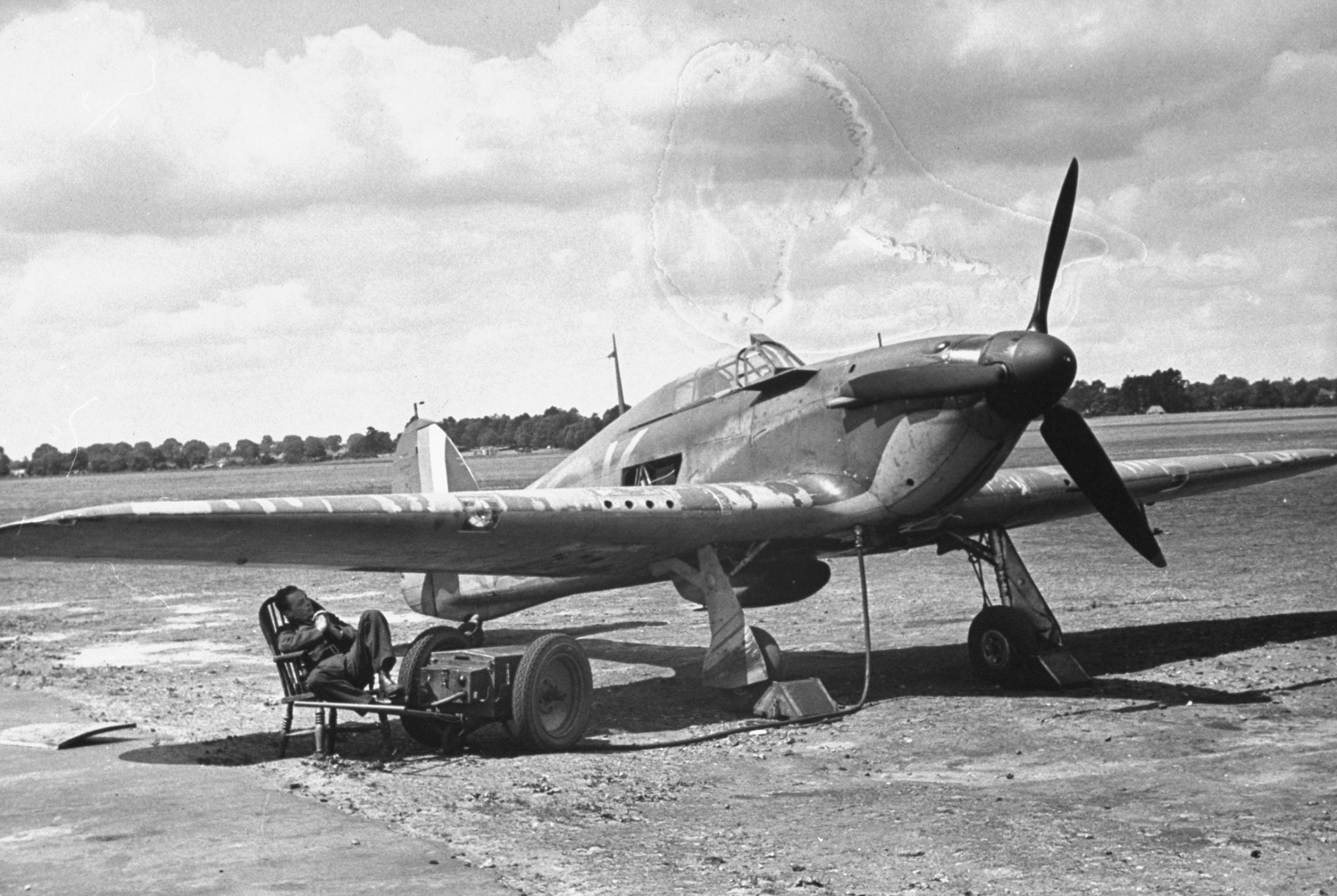 Between flights, member of ground crew sits in the shade of his plane's wing. Notice emergency starter apparatus at his feet, already hooked up, and four little holes in front edge of wing. Through these, four of plane's eight machine guns fire in unison.