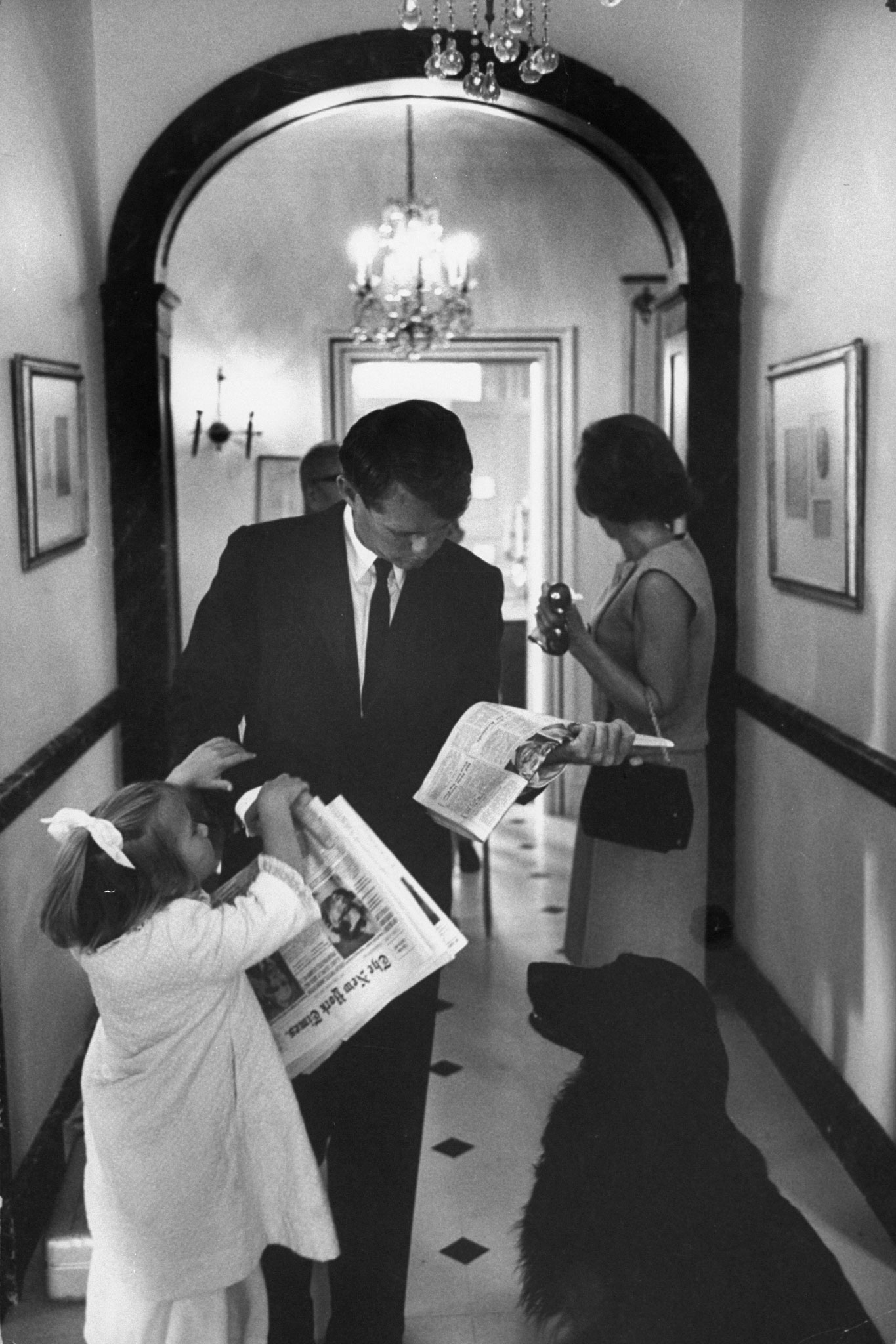 On his way to the office, Kennedy stands in hall at Hickory Hill with wife, Ethel, scanning the headlines while his daughter Kerry, 4, tugs for a goodbye kiss.