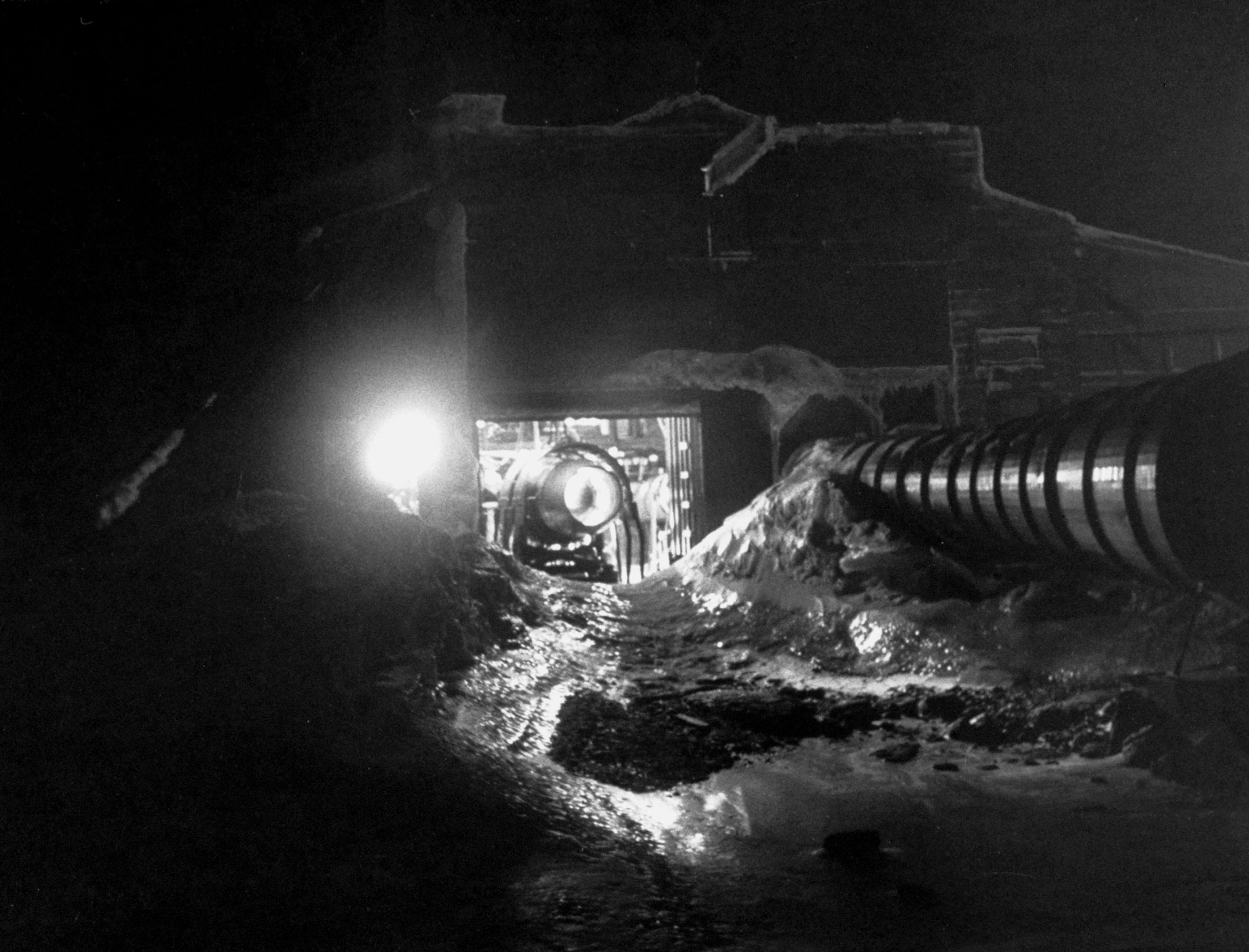 Jet test, carried on in an open-front steel hangar, is run day and night to measure and photograph ice which forms at the intake on the inside of mounted engine. Depression in snowbank is created by the engine's fiery blast. . . . When engine is turned off, water quickly refreezes rock-hard.