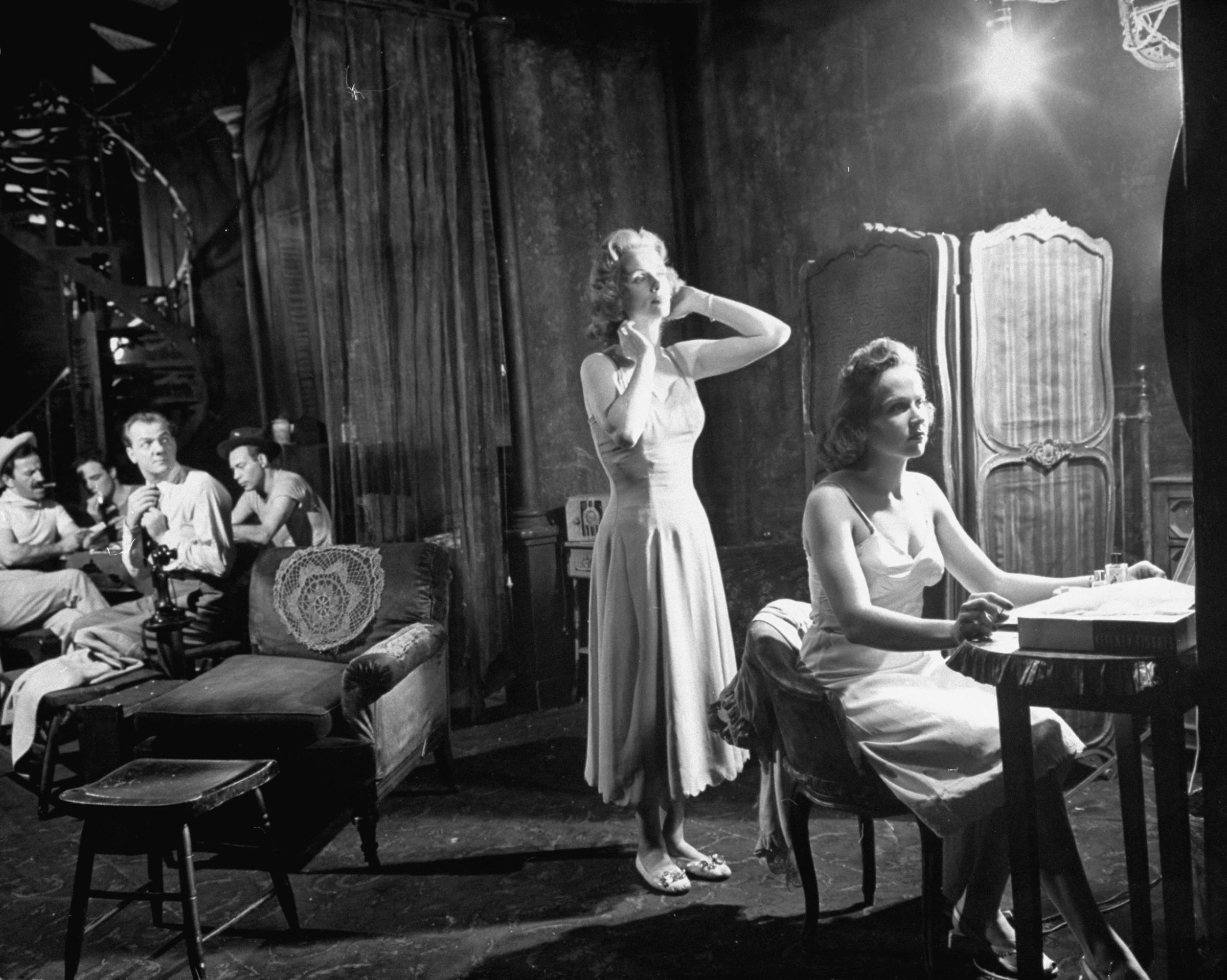 Blanche and Stella (Kim Hunter) undress in a bedroom which is divided from living room by partly closed curtains. Though Blanche complains about the noisy poker party which is going on in the adjoining room, she purposely stands so she can be seen by Mitch (Karl Malden, third from left).