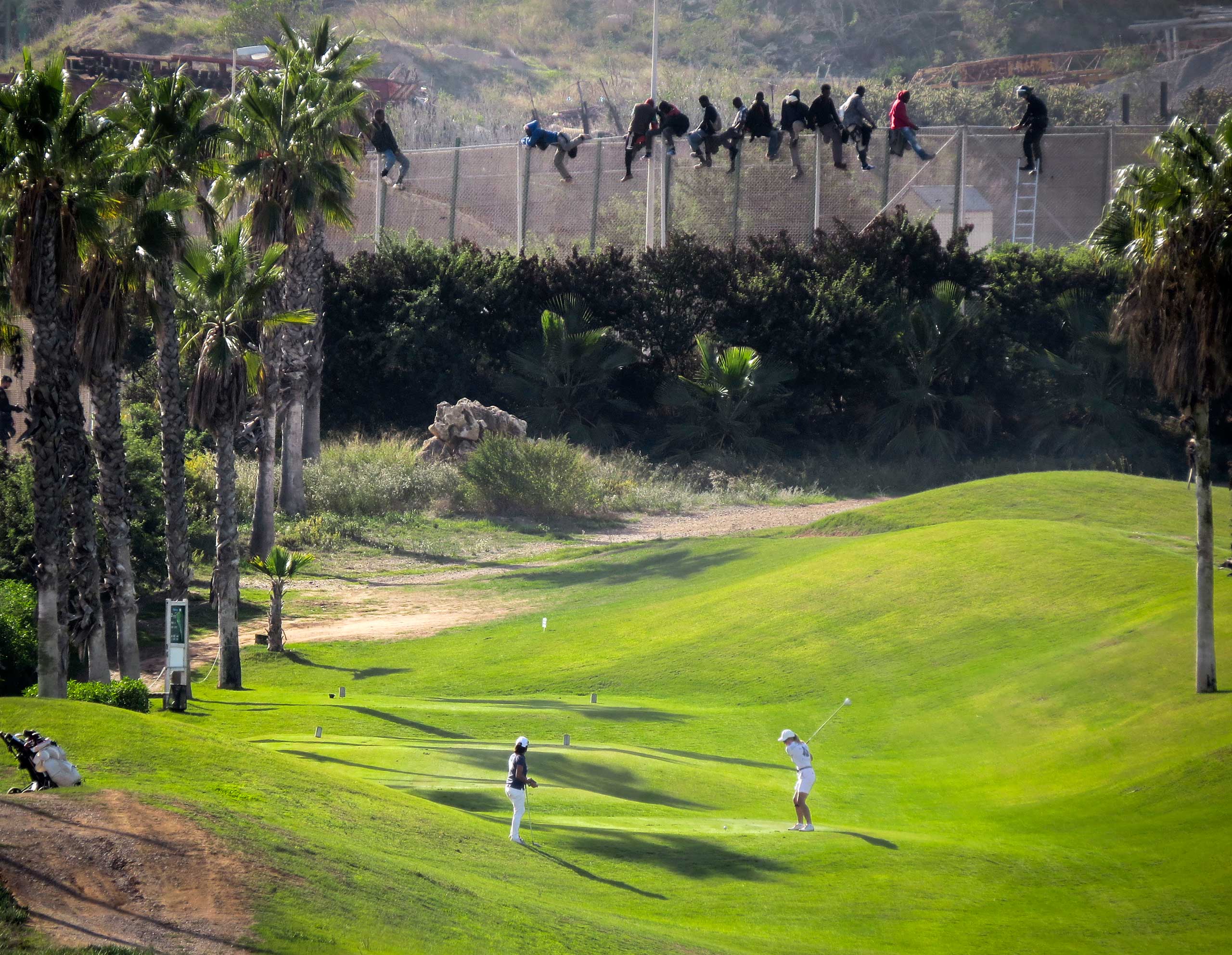 A golfer hits a tee shot as African migrants sit atop a border fence during an attempt to cross into Spanish territories between Morocco and Spain's north African enclave of Melilla, Oct. 22, 2014.