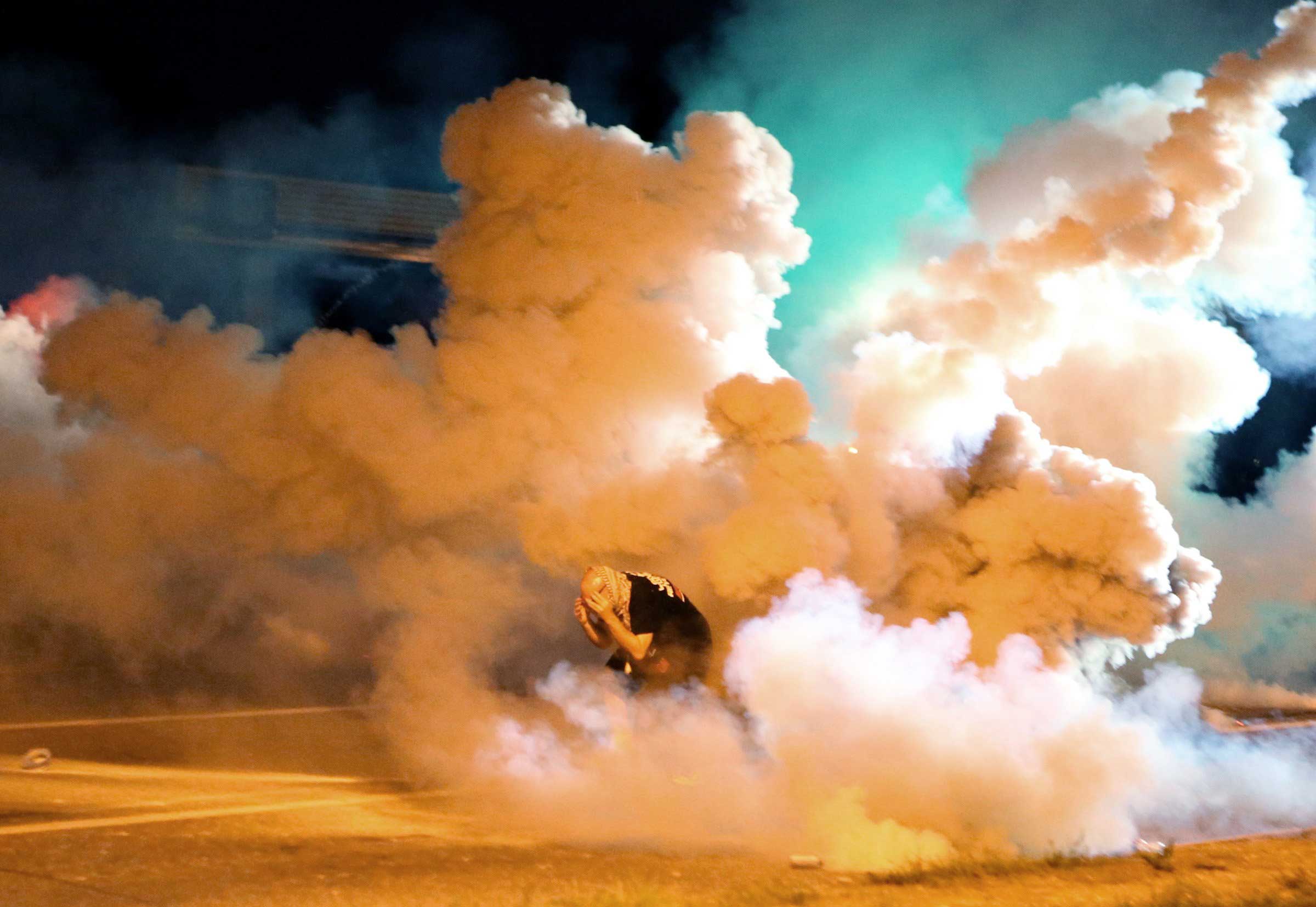 Aug. 13, 2014. A protester takes shelter from smoke billowing around him in Freguson, Mo. Protests in the St. Louis suburb rocked by racial unrest since a white police officer shot an unarmed black teenager to death turned violent Wednesday night, with some people lobbing Molotov cocktails and other objects at police who responded with smoke bombs and tear gas to disperse the crowd.