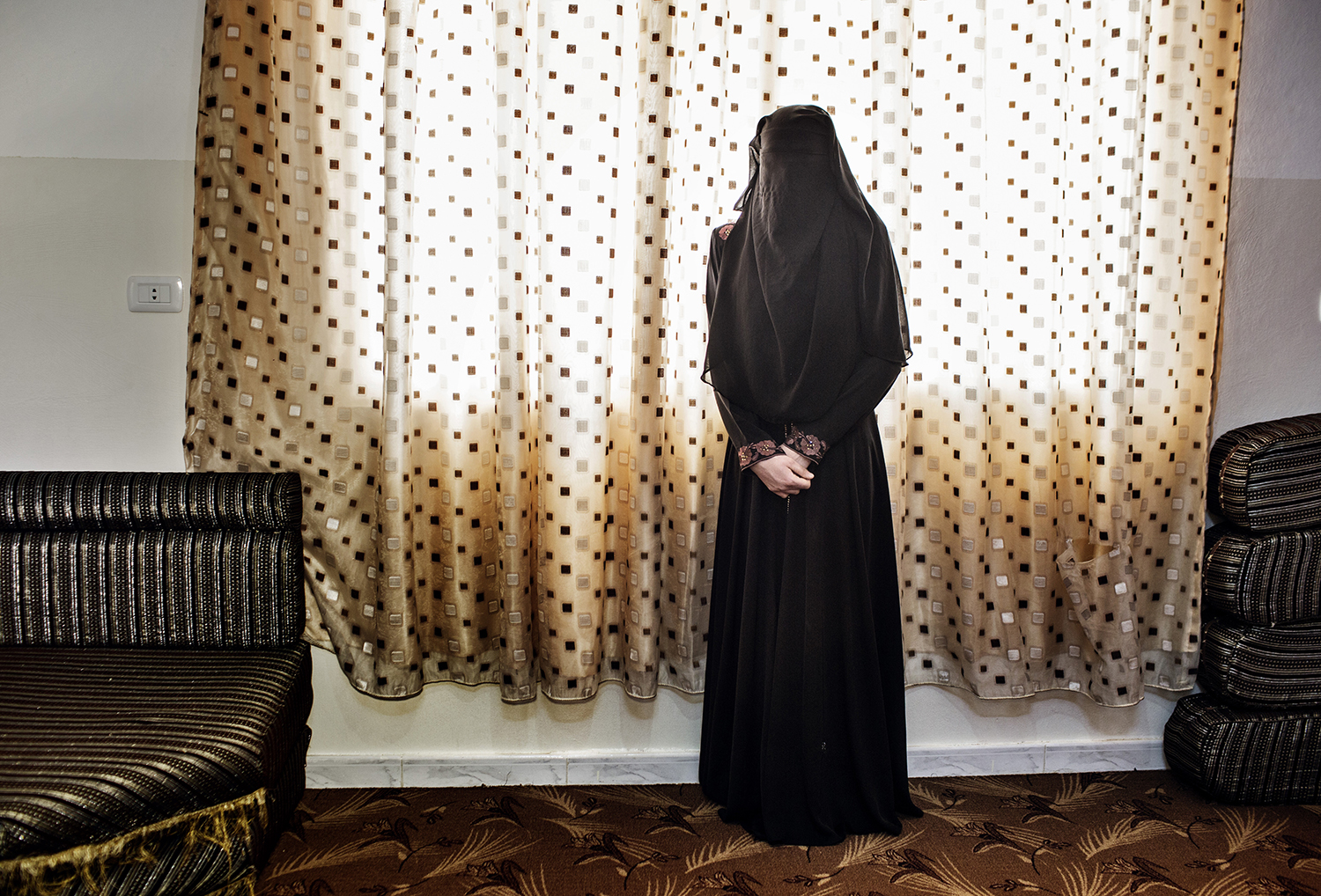 TOMORROW THERE WILL BE APRICOTS:2014, M, 15, (previously pictured w her sister) stands alone in the niqab she recently started wearing. Her 17 year old sister has just married, leaving her alone in the house with her widowed mother and four siblings. (they live in a "martyrs wive" building). The marriage proposals are already coming to her. She had dreamt of finishing high school and becoming a teacher. She says that while early marriage was common in Deraa, at least girls were allowed by their families to walk freely in their community and go to school. Here, she feels she is slowly dying indoors, day in and day out.Their mother is fairly conservative, and does not mind the rules of the "martyrs wives building". She says the are only worried about our safety and reputation and we are grateful for that. The Syrian ÒWives of MartyrsÓ are struggling to find a sense of normalcy in the dusty Jordanian border town of Ramtha, painfully close to the home and life they once had in Deraa. The burdens of violence are present in their scant belongings, heavy mementos to remind themselves of those they lost in the war. Digital era lockets: cherished cell phone images of dead fathers, husbands, and brothers lost to SyriaÕs bloody uprising. ÒTomorrow there will be apricotsÓ is a popular proverb in the Levant, which means, ÒTomorrow never comes.Ó I first met many of these women in 2012 when their husbands and fathers were still alive, fighting for the FSA. The photos explore intimacies of everyday life of four families headed by wives who have lost their fighter husbands to the civil war. They show the quiet celebrations and daydreams of the wives and daughters of martyrs who now live in a building donated by a Qatari patron, despite traditions that frown upon displays of joy for single women, who are already caught in a vicious cycle of poverty, isolation, and anxiety.