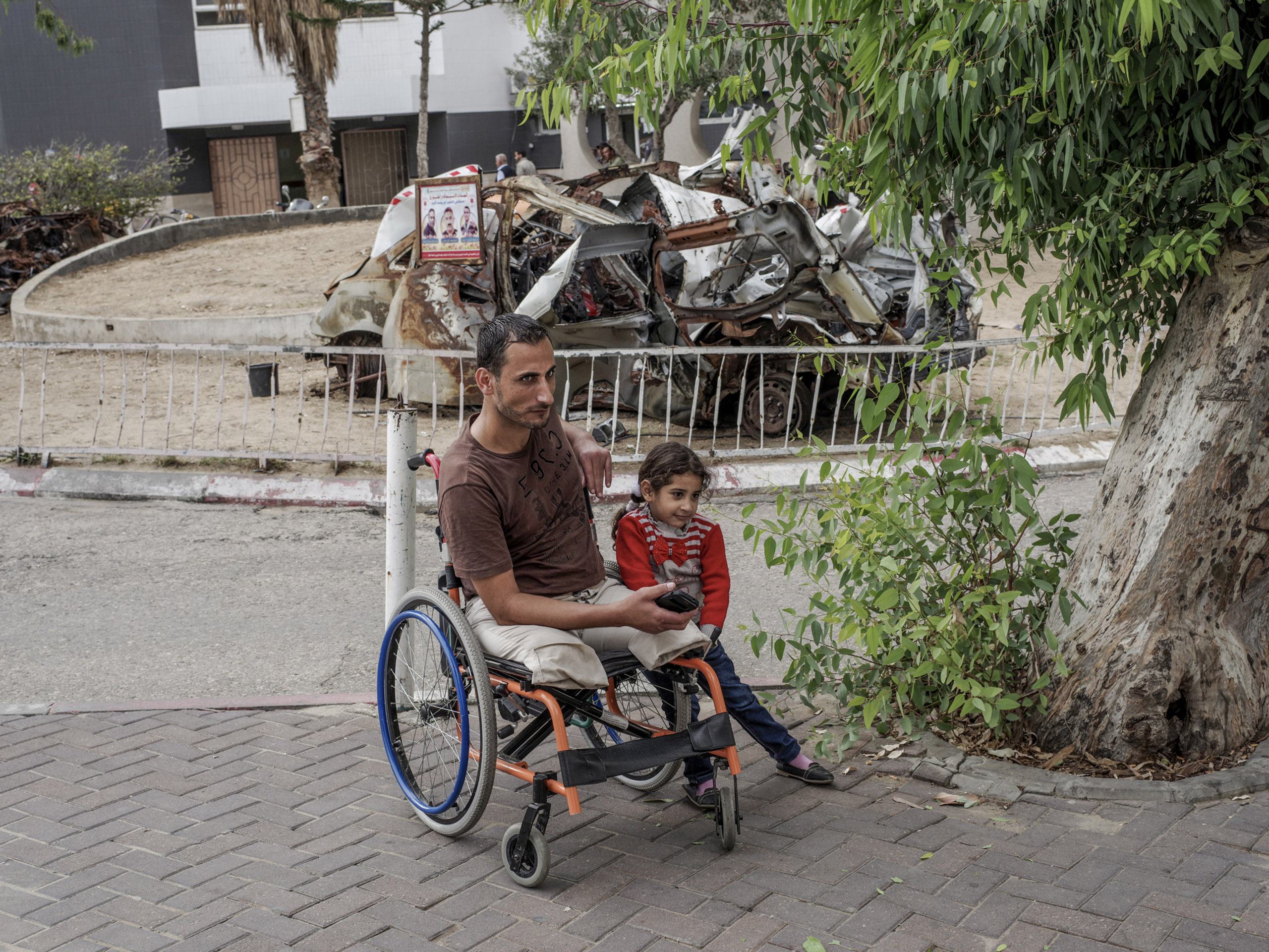 Achmed Hassan and his daughter. Hassan lost both his legs in 2008 and is still in recovery.  The destroyed vehicle in the background is an ambulance destroyed in during Operation Protective Edge.