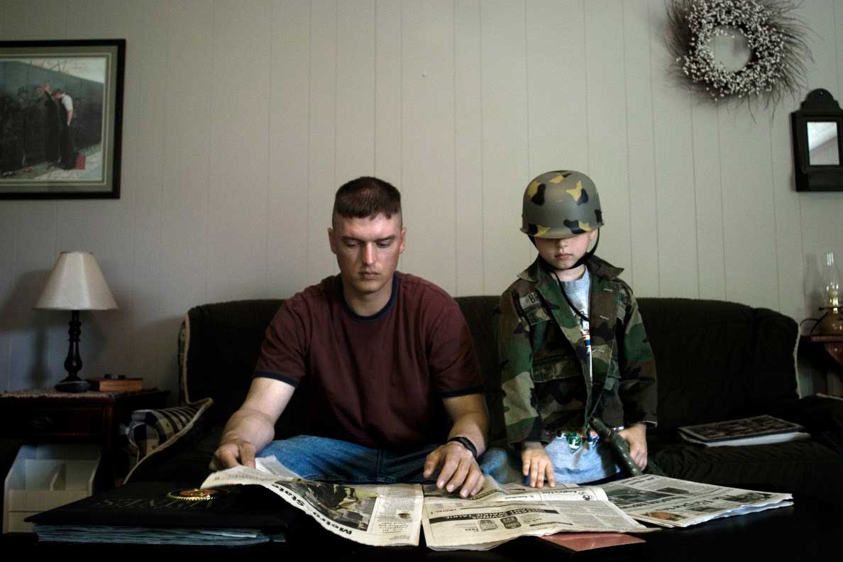 Staff Sergeant Travis Brill.Andrea Bruce, 2006. Columbus, Ohio.
                               This scene looks unremarkable at first. A quiet morning Sergeant Travis Brill, a Marine just home from Iraq, reads the paper. But two things stood out to me. First, the desperate desire of his family to connect and cross an expanse of experiences--to see what he sees. His son dresses like him, mimics him. His wife decorates the house with a print from the Vietnam Memorial. Everyone tries to understand, to share in the pain or grief or courage or rage, while Travis remains far away, still in the war. This subtle moment also shows the changing relationship we have to war in a lifetime. It starts as a toy-gun spectacle, changes to real life, the now, Travis’ war. (He deployed again in 2010). And then, remembrance. Iraq changed the people experiencing the war more than they could have imagined, guiding and afflicting them in ways many are still struggling to understand. At the time of this photo, in 2006, Lima took more casualties than any U.S. company in Iraq, losing 23 Marines killed in action.