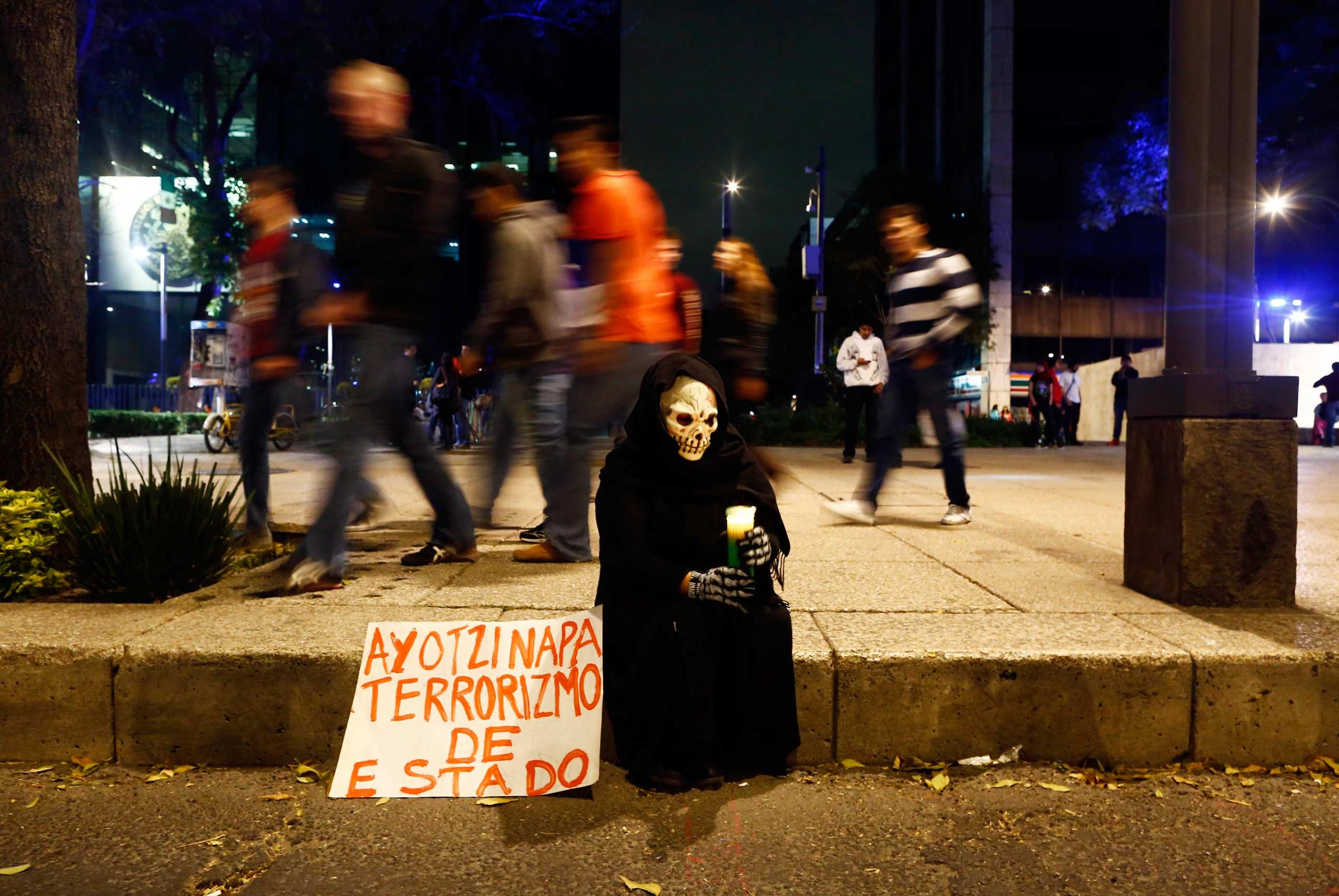 A person in a disguise sits on a sidewalk holding a candle during a protest denouncing the apparent massacre of 43 trainee teachers in Mexico City